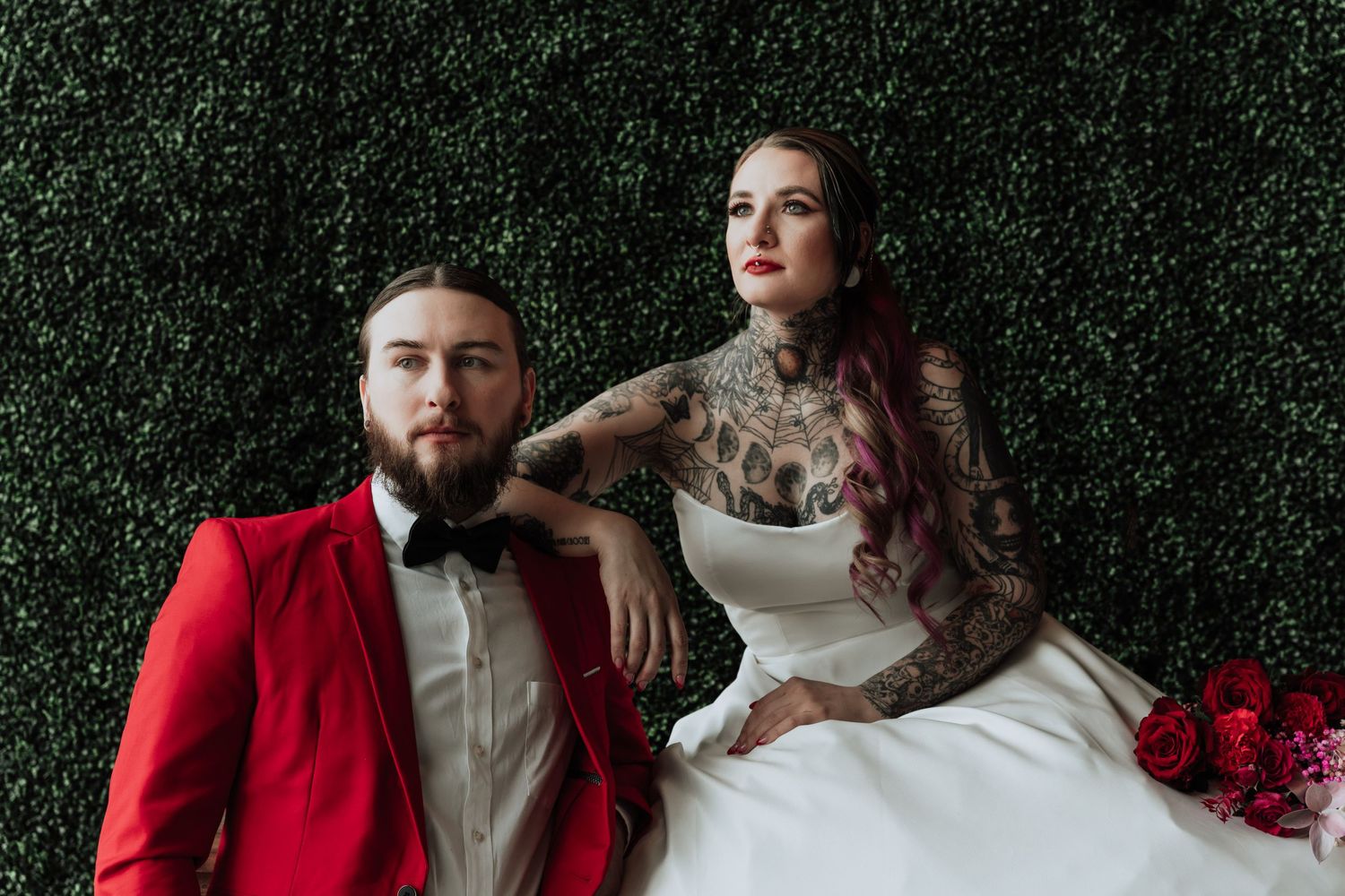 Wedding couple in red suit and white dress pose against hedge wall with dramatic styling.