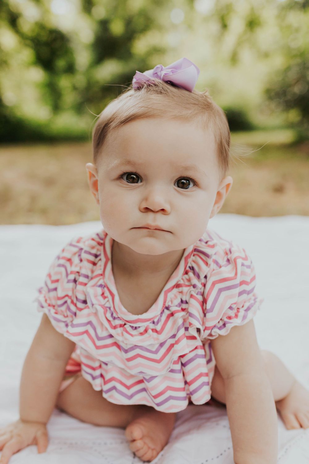 Baby in pink chevron dress posing on white blanket during outdoor portrait session.