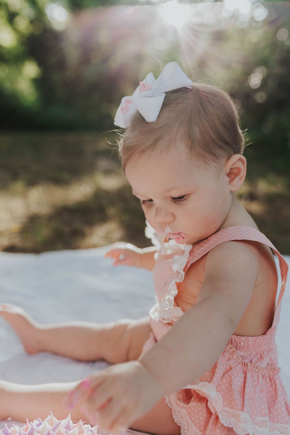 Baby with white bow accessory wearing pink outfit during outdoor photo session.