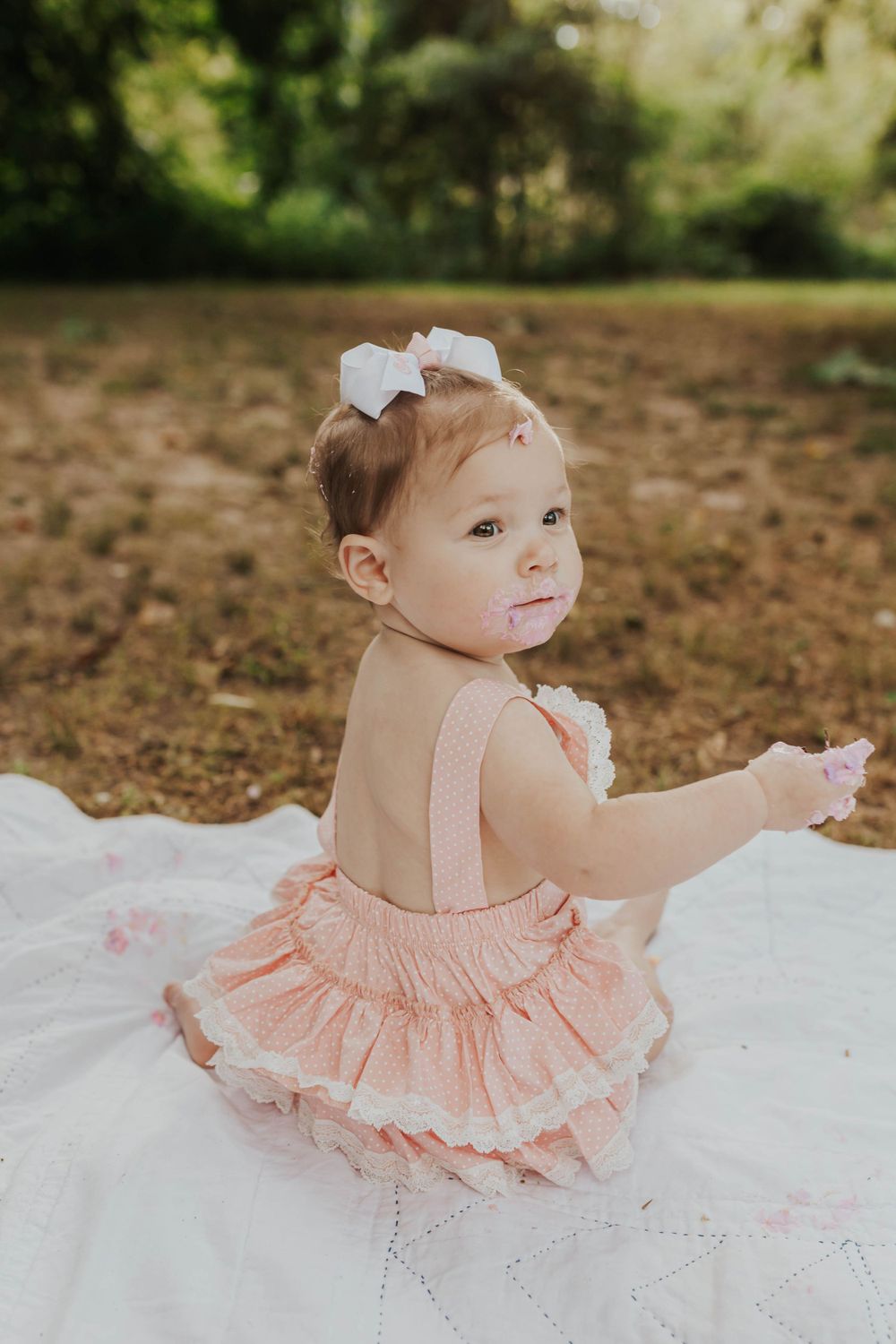 Baby in a peach-colored ruffled dress plays on a white blanket outdoors during a photo session.