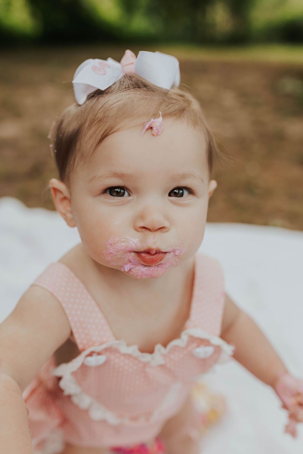 Baby wearing a peach dress and white bow smiles with cake frosting on their face.