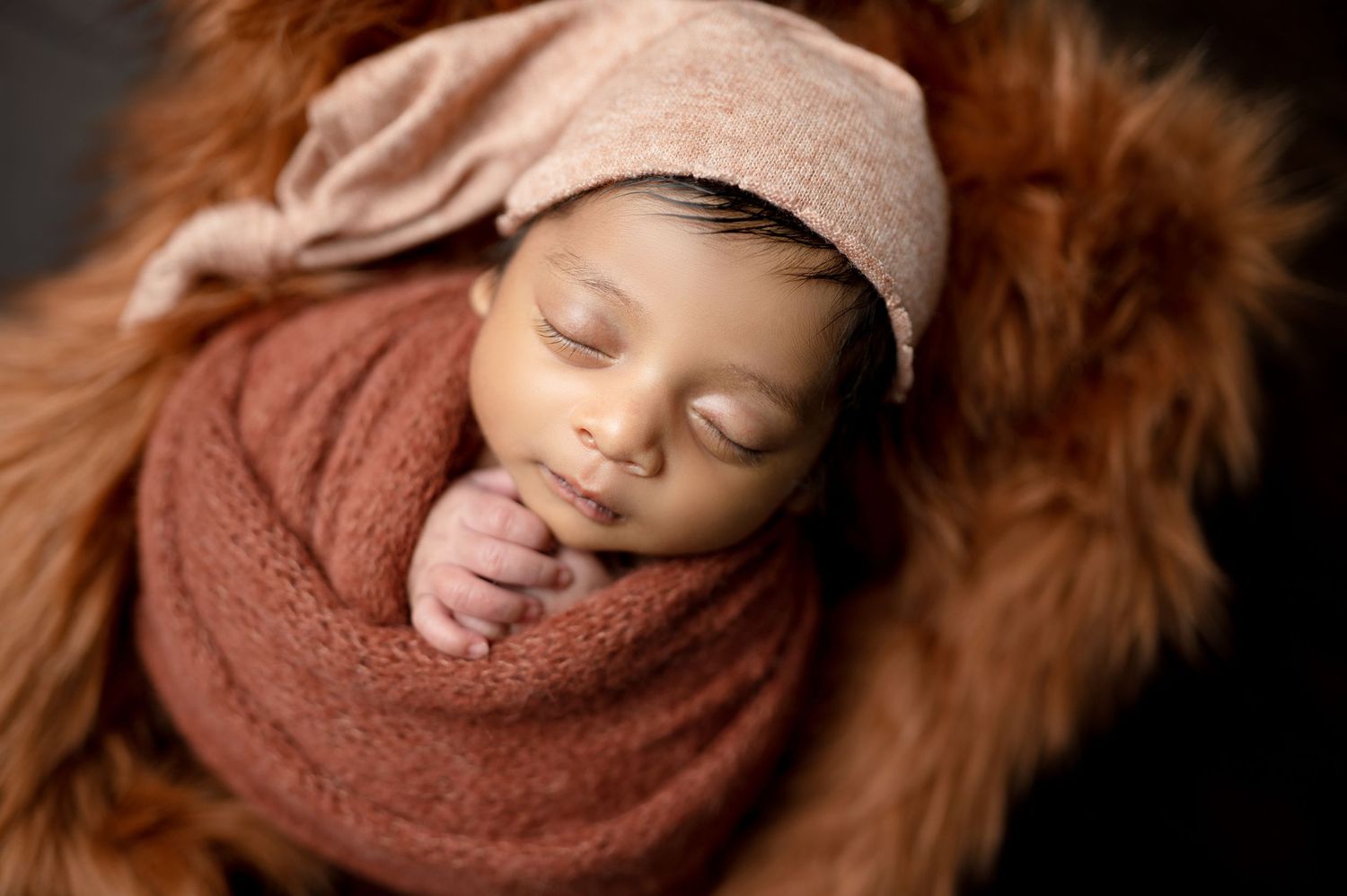 Sleeping newborn wrapped in brown swaddle on fur blanket in artistic portrait series.
