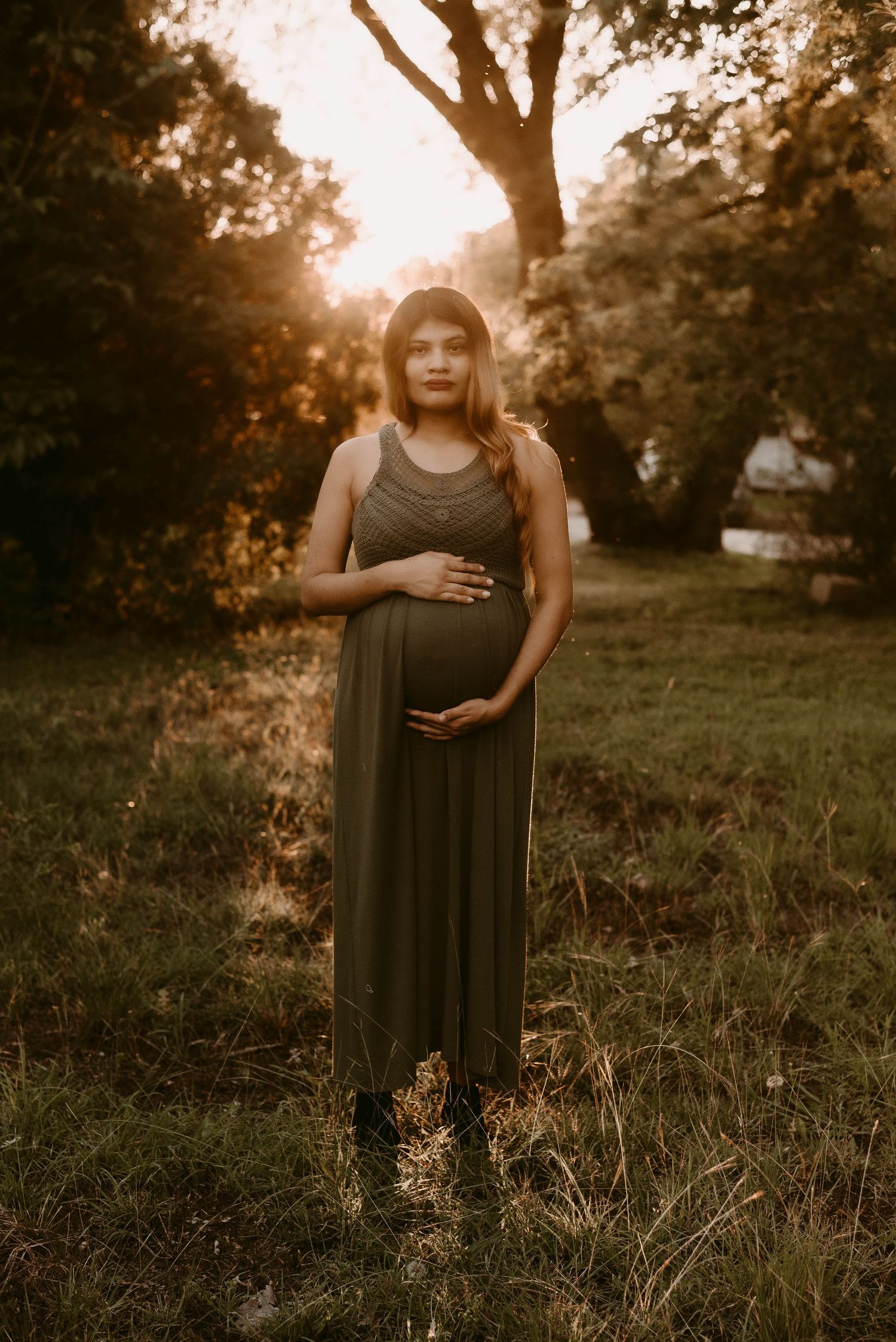 Maternity portrait in maxi dress against backlit sunset trees.
