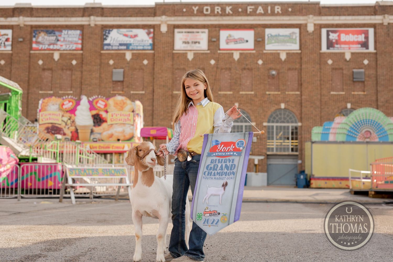 A person walks alongside their white and brown dog at a fairground with carnival rides visible in the background.