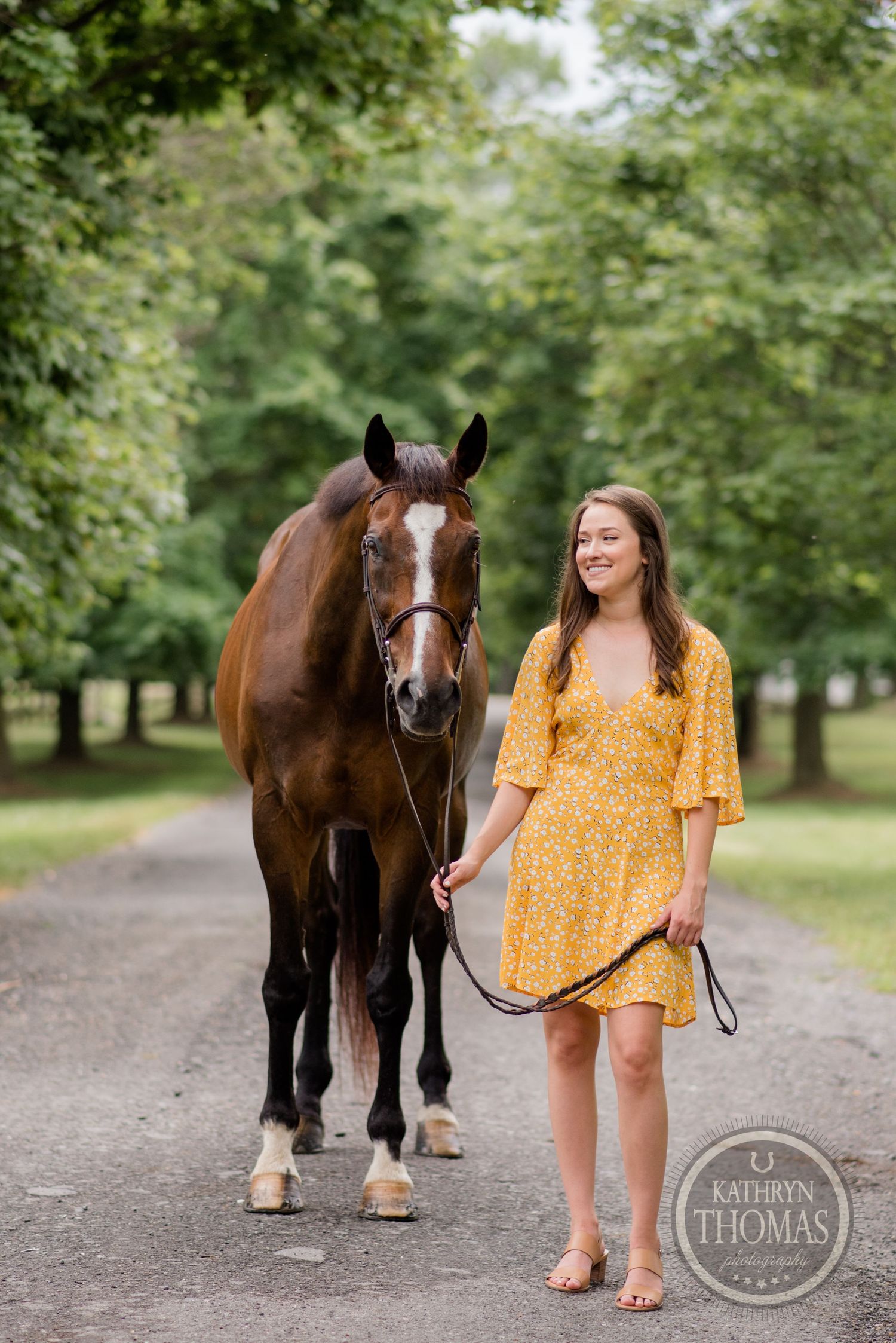 A bay horse and companion in a yellow polka dot dress stand together on a peaceful wooded path.
