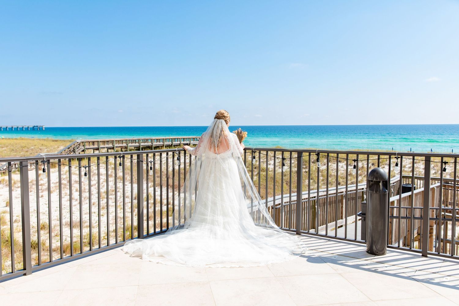 A bride in a flowing white gown and veil stands at a beach boardwalk overlooking the ocean.