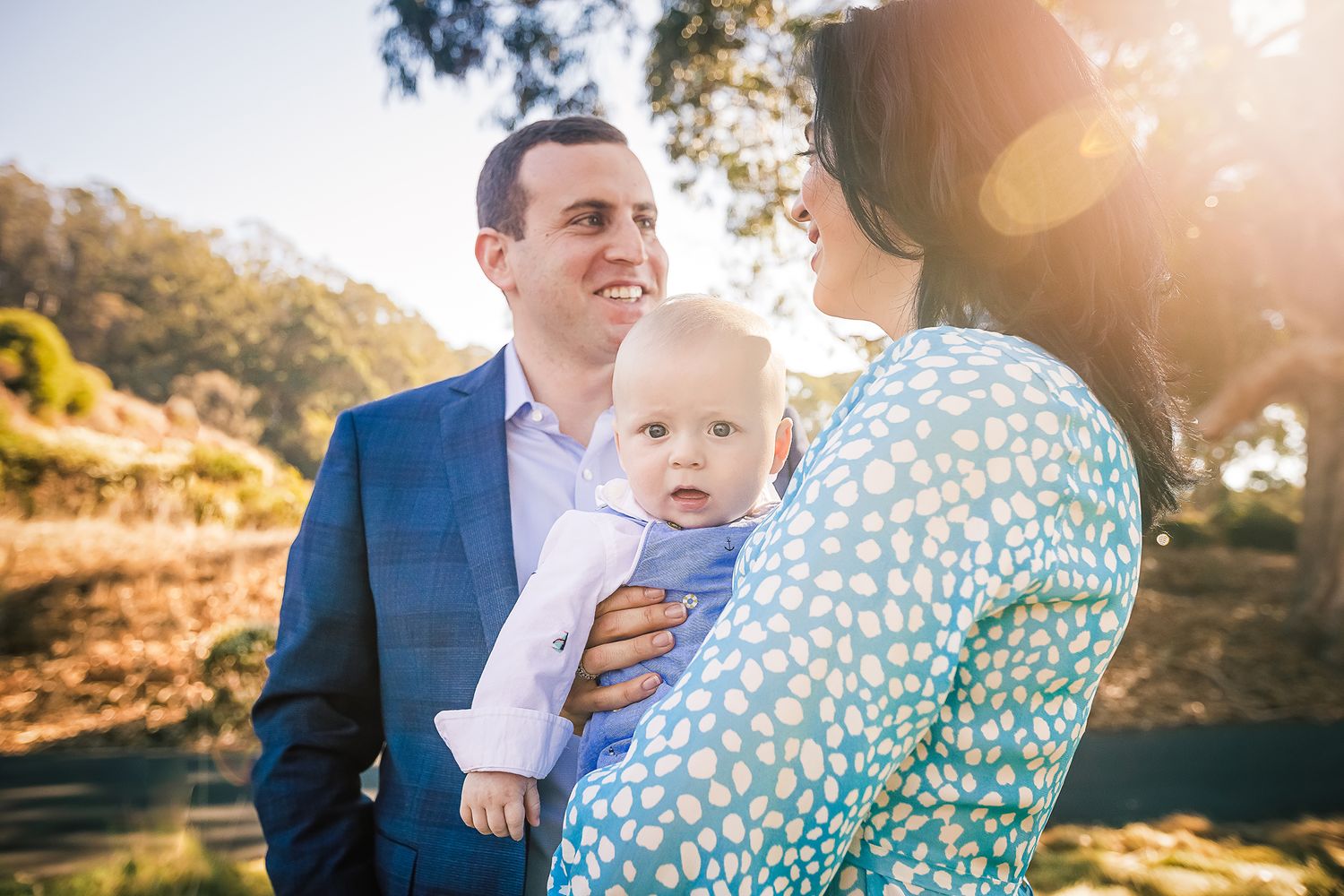 On a sunny day mom wearing a light blue dress and dad wearing a jacket smiling at each other happily holding their baby