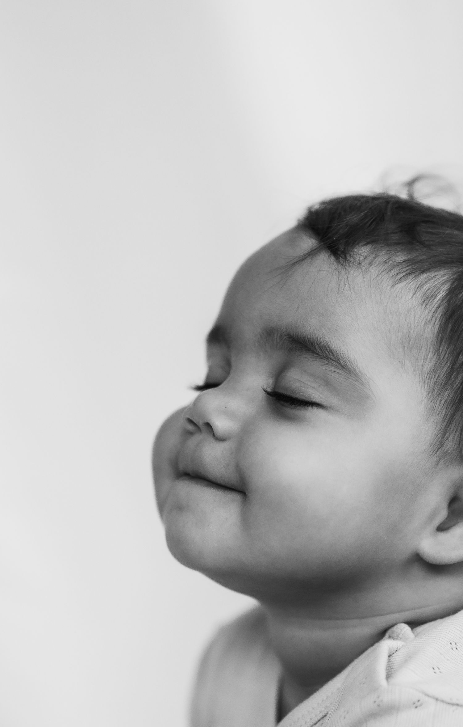 Close up black and white image of a toddler smiling cheekily with her eyes closed.