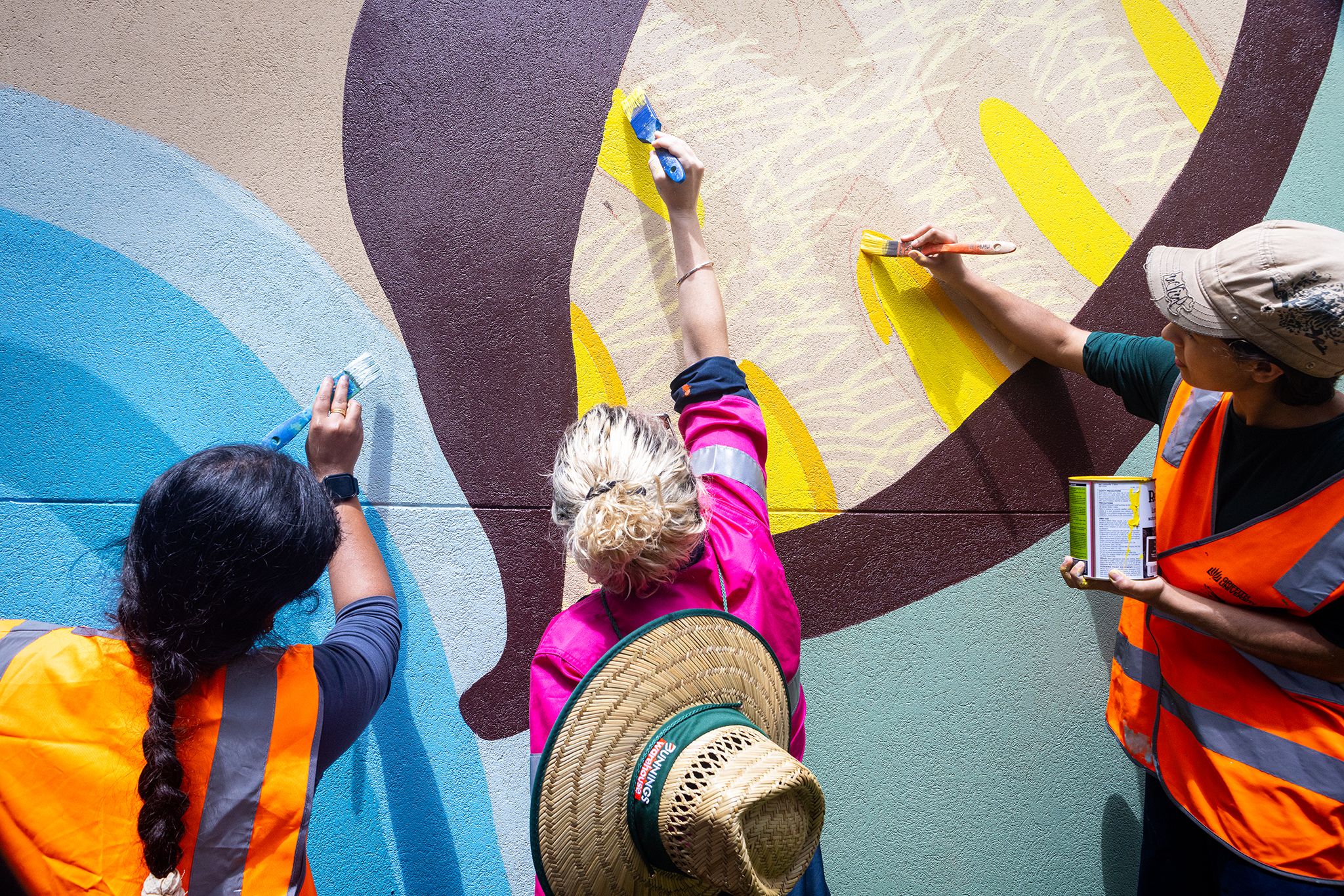 Three women paint a mural on a wall with bright coloured paint.