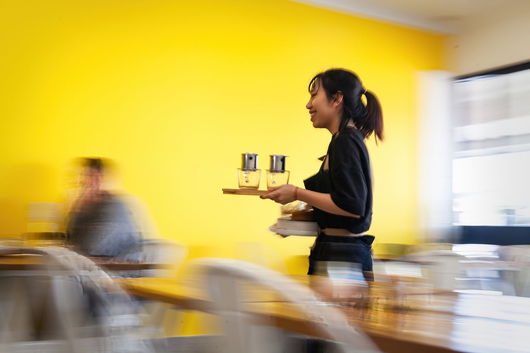 A young Asian waitress carries Vietnamese iced coffee to a customer in a cafe. In the background is a yellow wall.