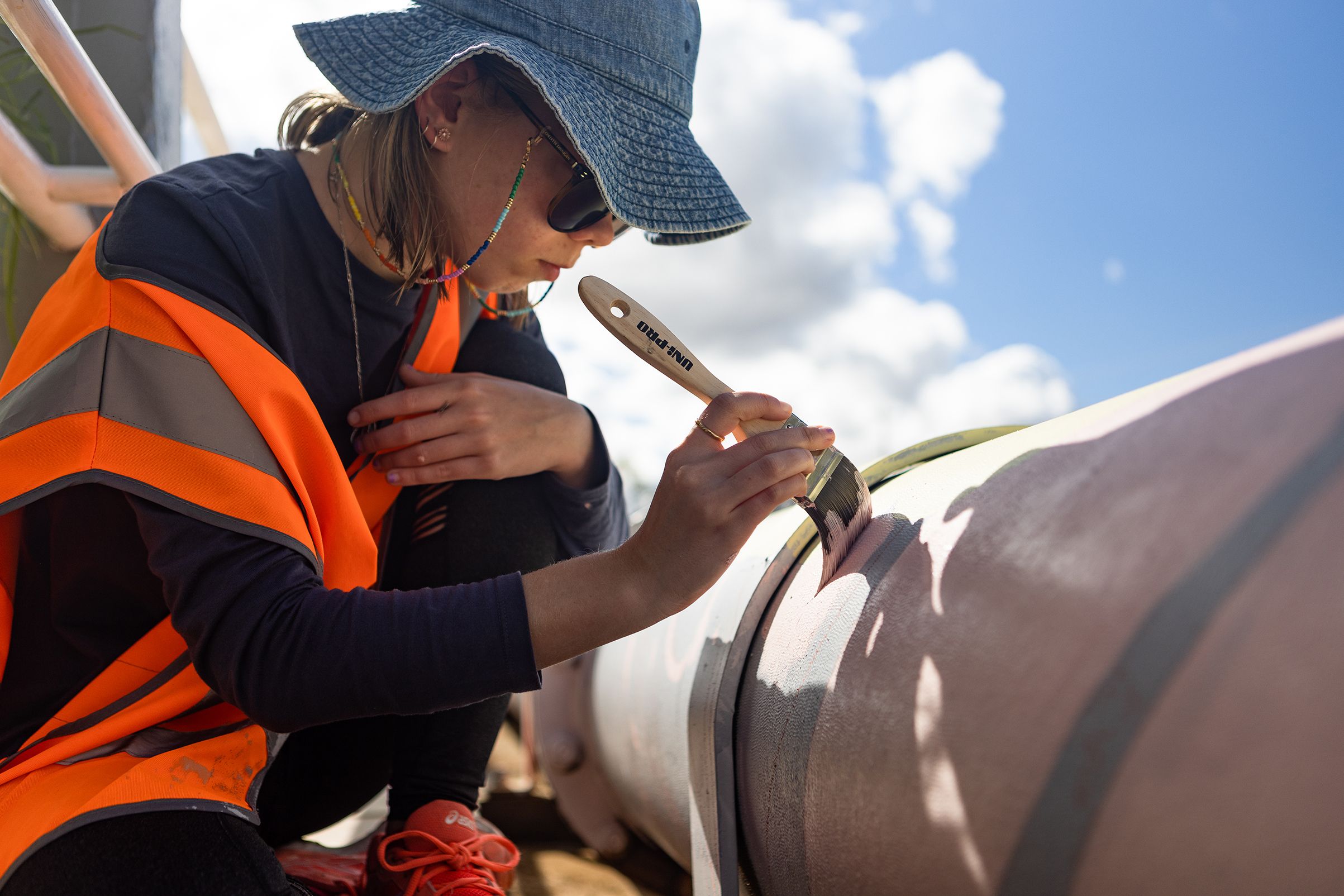 A young woman holding a paint brush paints a mural onto a pipe outdoors.
