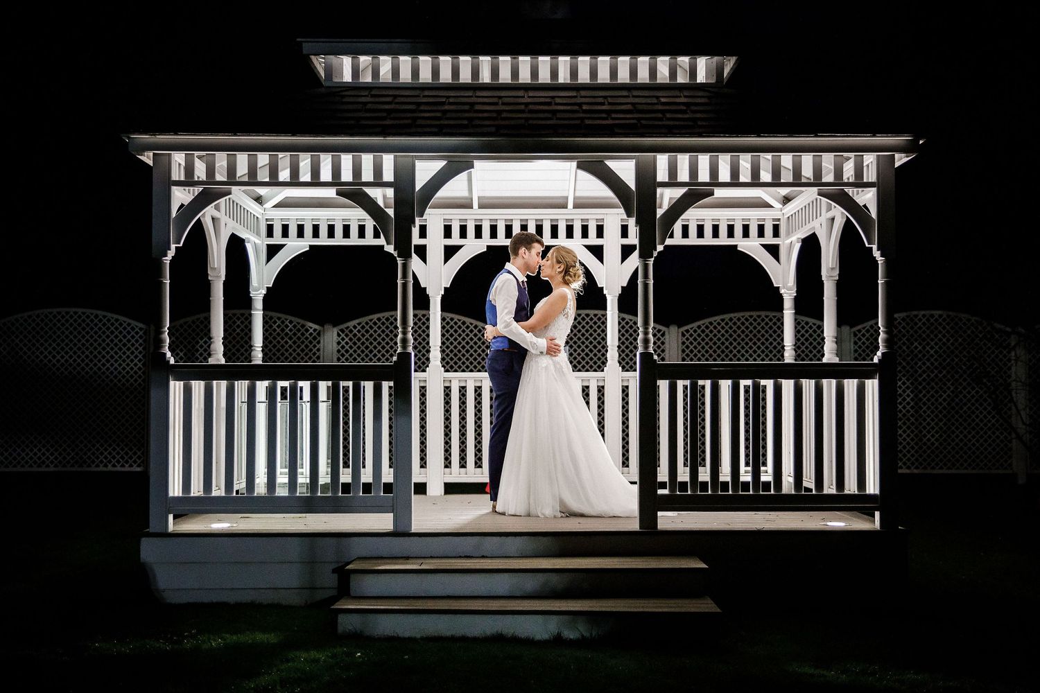 Wedding couple shares a romantic moment under an elegant white gazebo at night during outdoor reception.