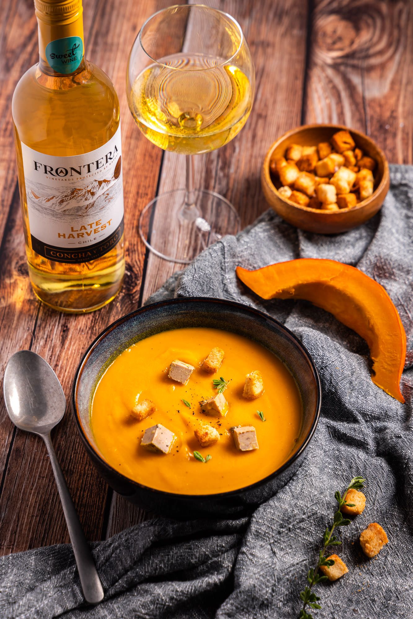 Creamy pumpkin soup with croutons served alongside wine bottles on wooden table.