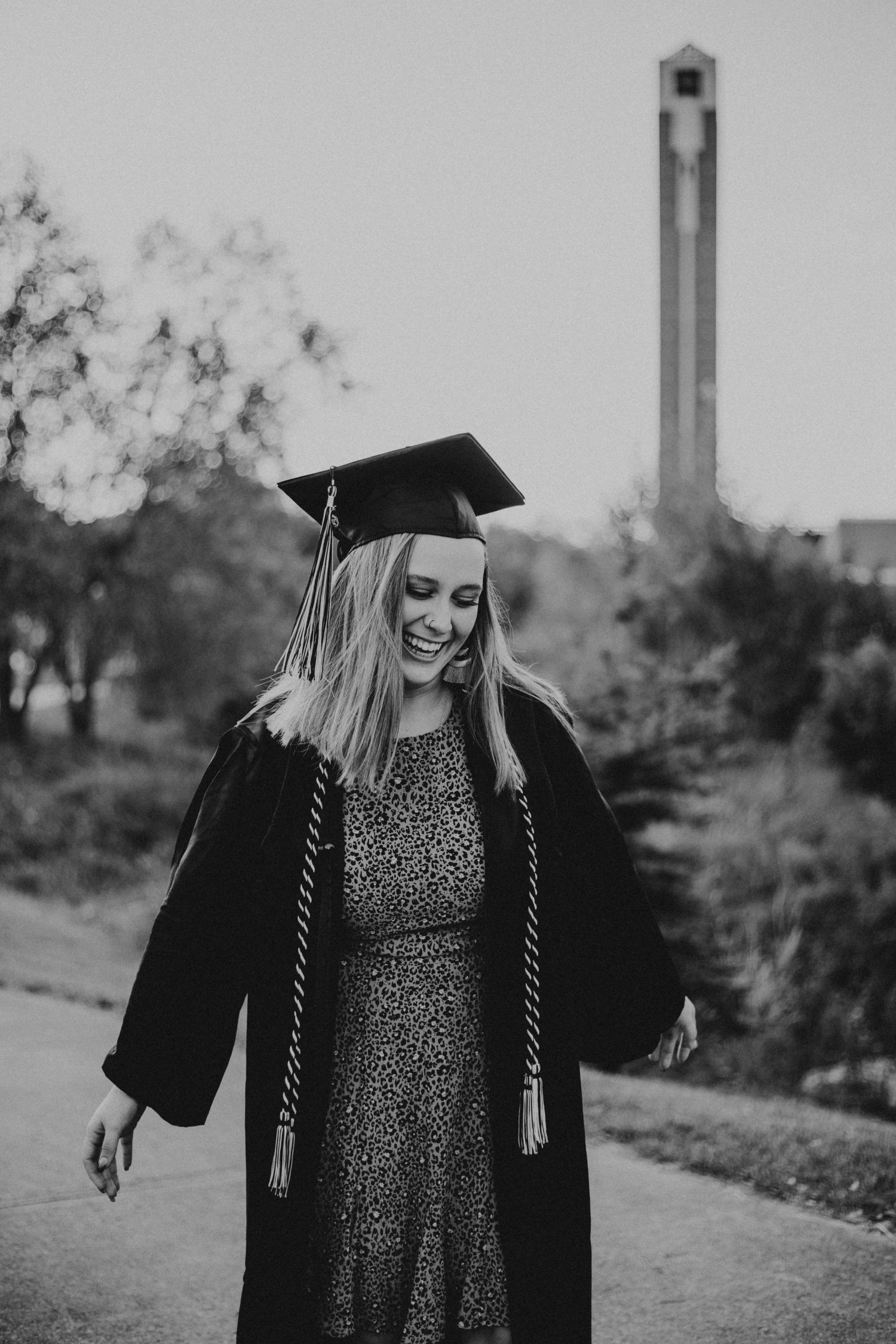 Graduate in sparkly dress and black gown smiling with bell tower in background.