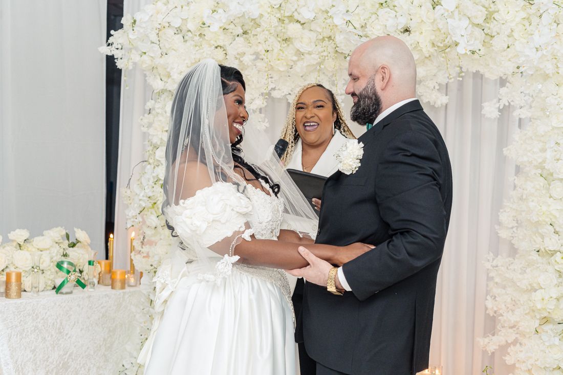 Wedding ceremony moment with couple and officiant in front of white floral wall.