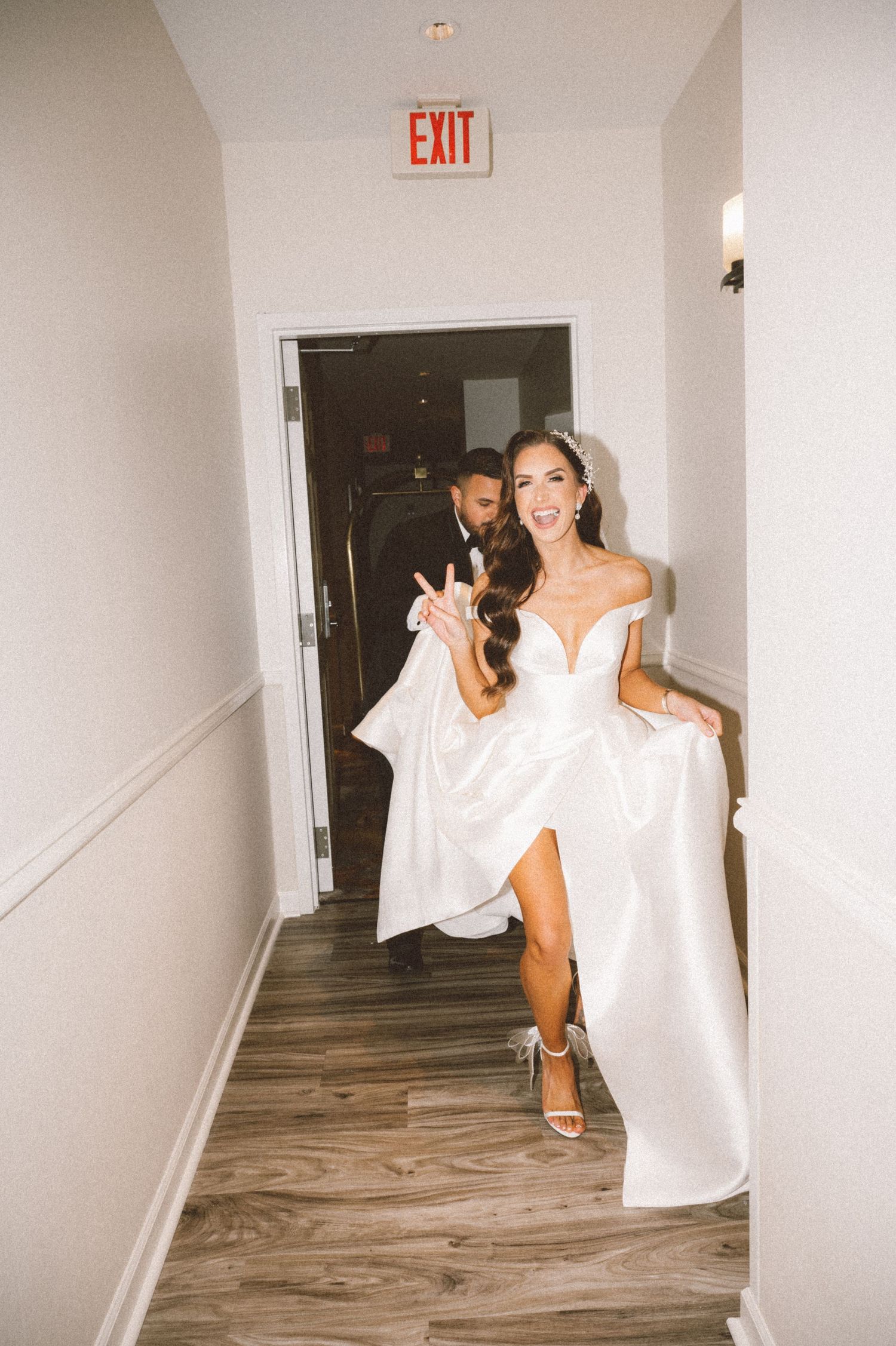 Joyful bride in white slit dress poses in hallway under exit sign.