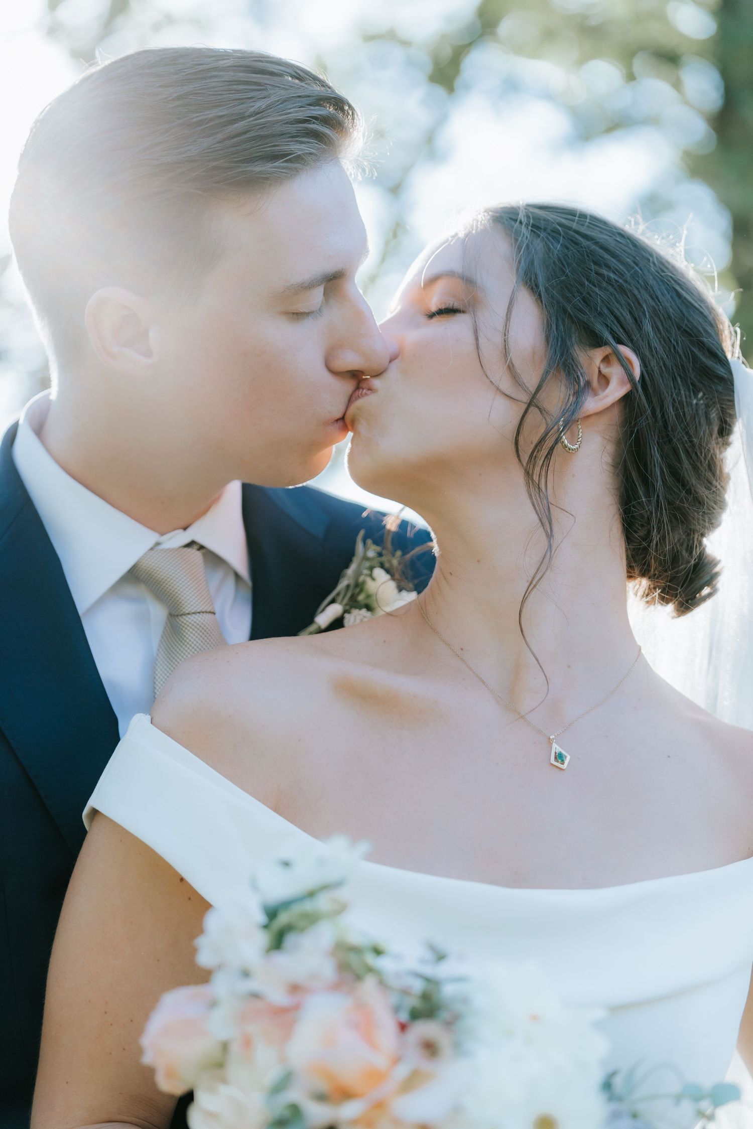 Tender wedding day kiss captured in soft natural lighting with bridal bouquet.
