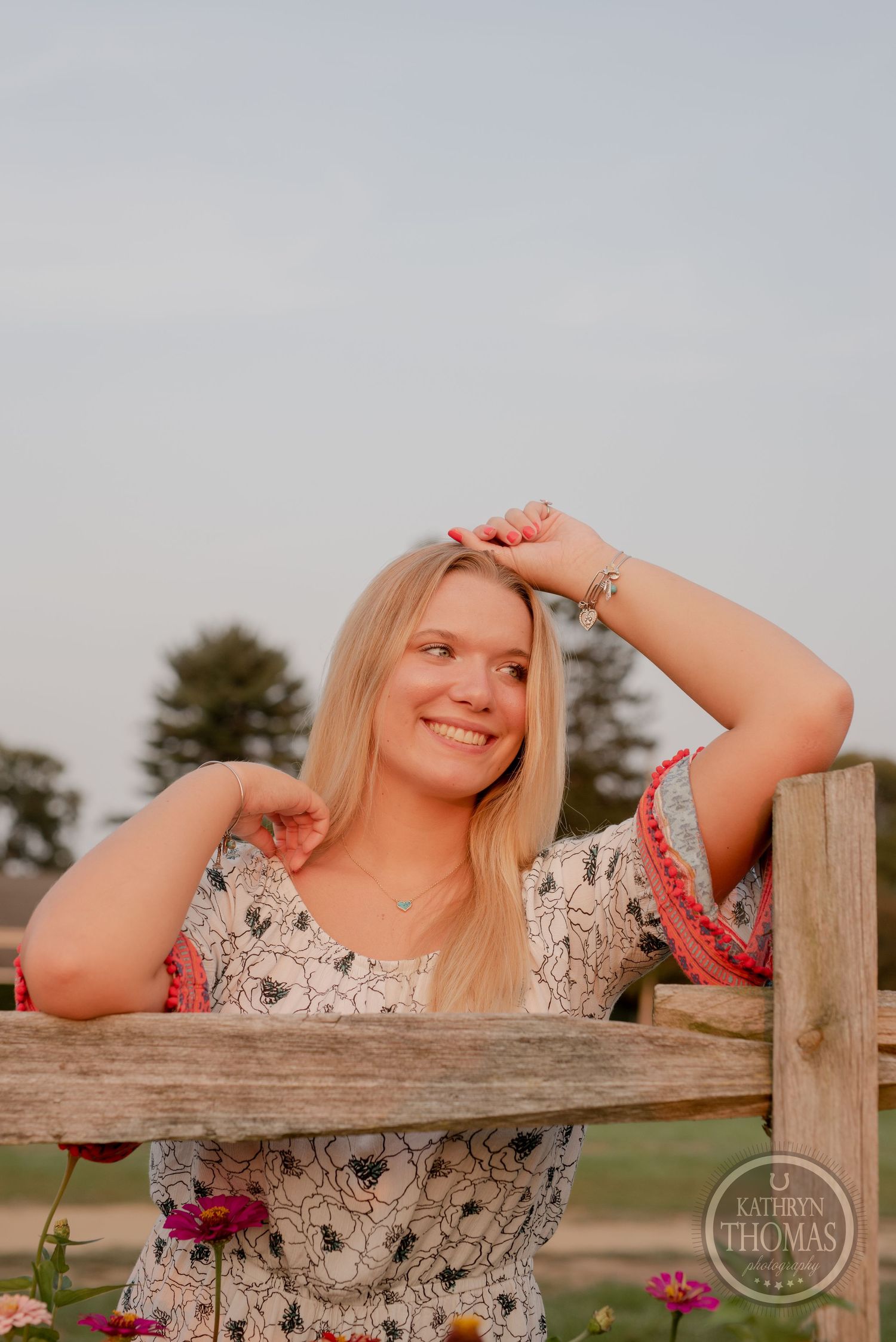 A woman leans on a wooden fence with flowers blooming nearby at sunset.