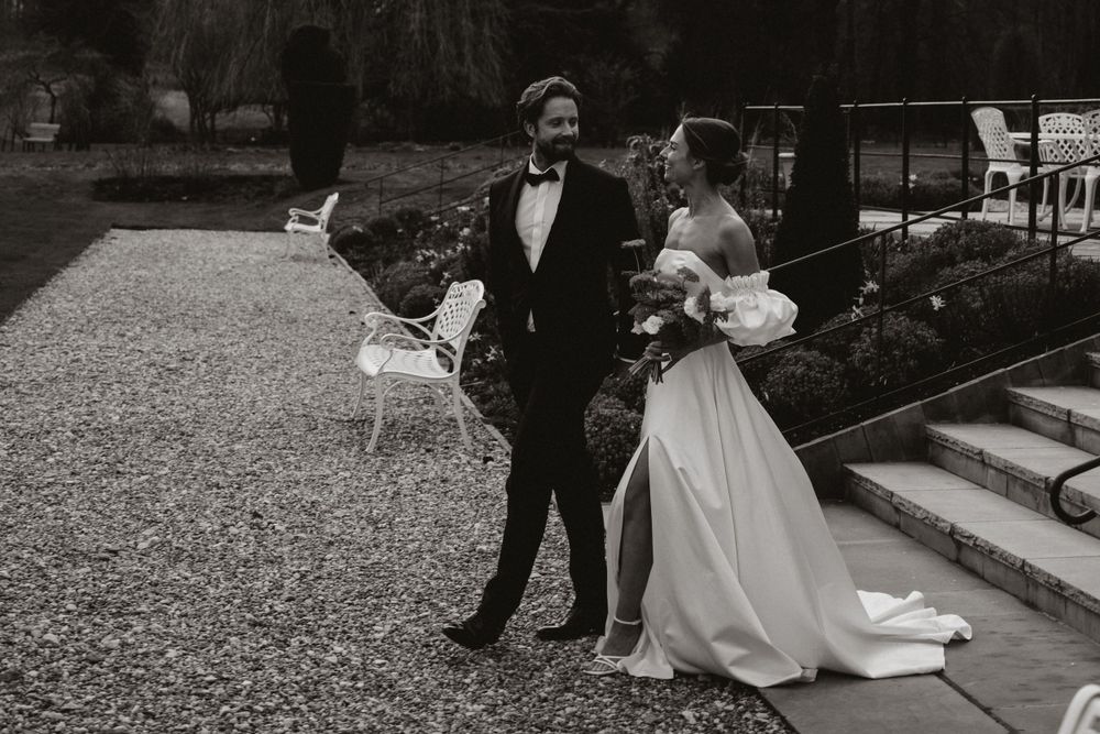 A black and white photo of a couple in wedding attire standing on a gravel path in a garden setting.