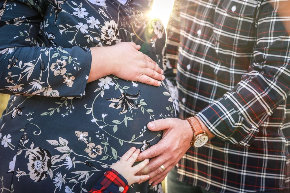 a trio of hands touch a woman's pregnant belly with the sun shining between them