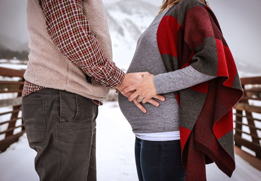 A couple holds their hands on her baby bump while standing on a bridge in the snow in Crested Butte Colorado