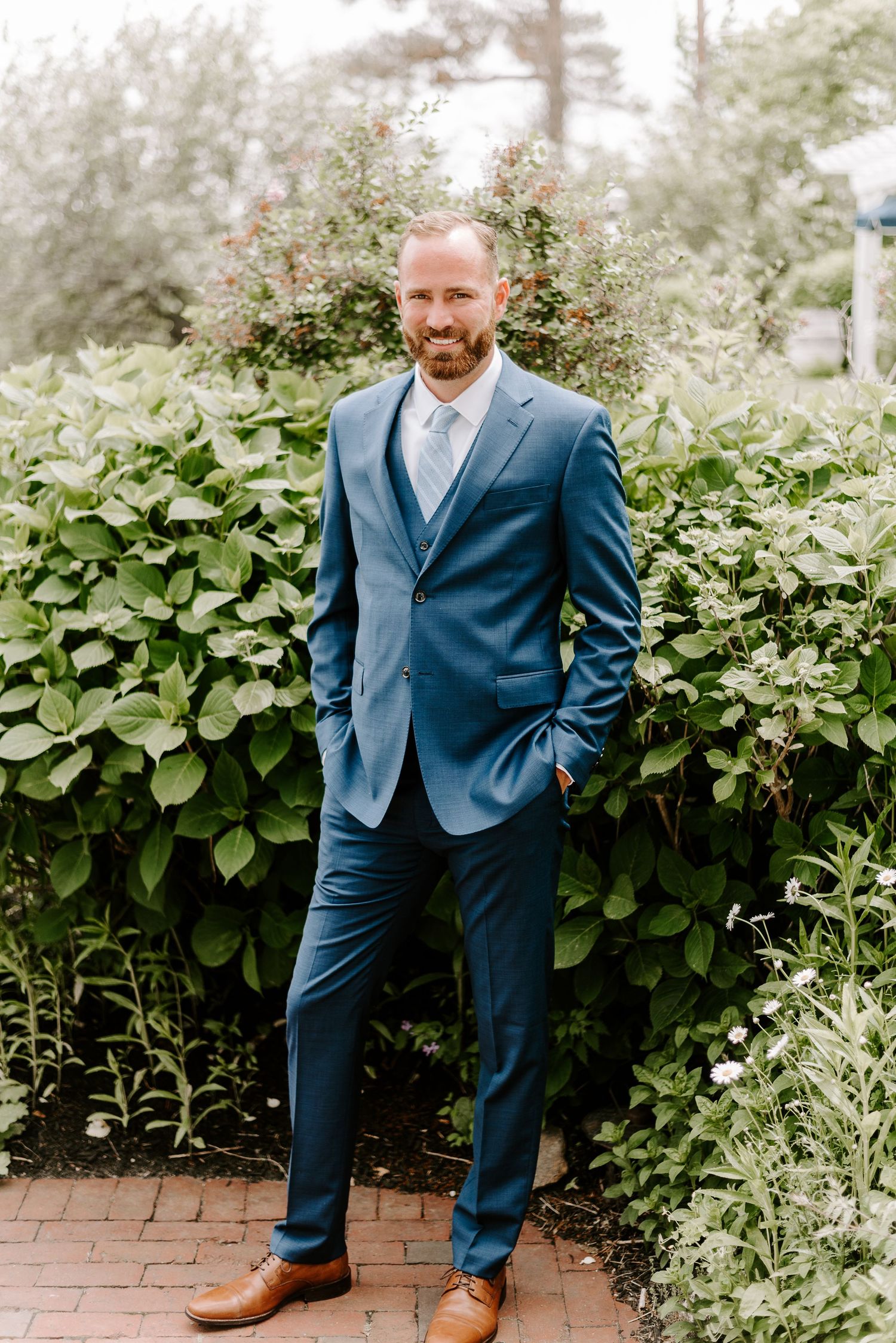 Wedding party member in navy suit poses against garden greenery backdrop.