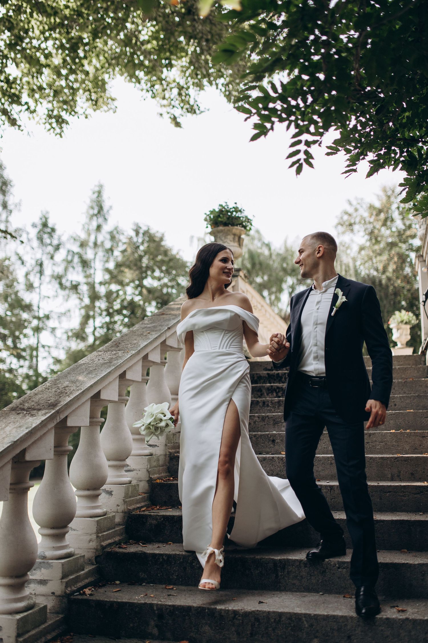 A couple in wedding attire poses on stone steps in an elegant garden setting.