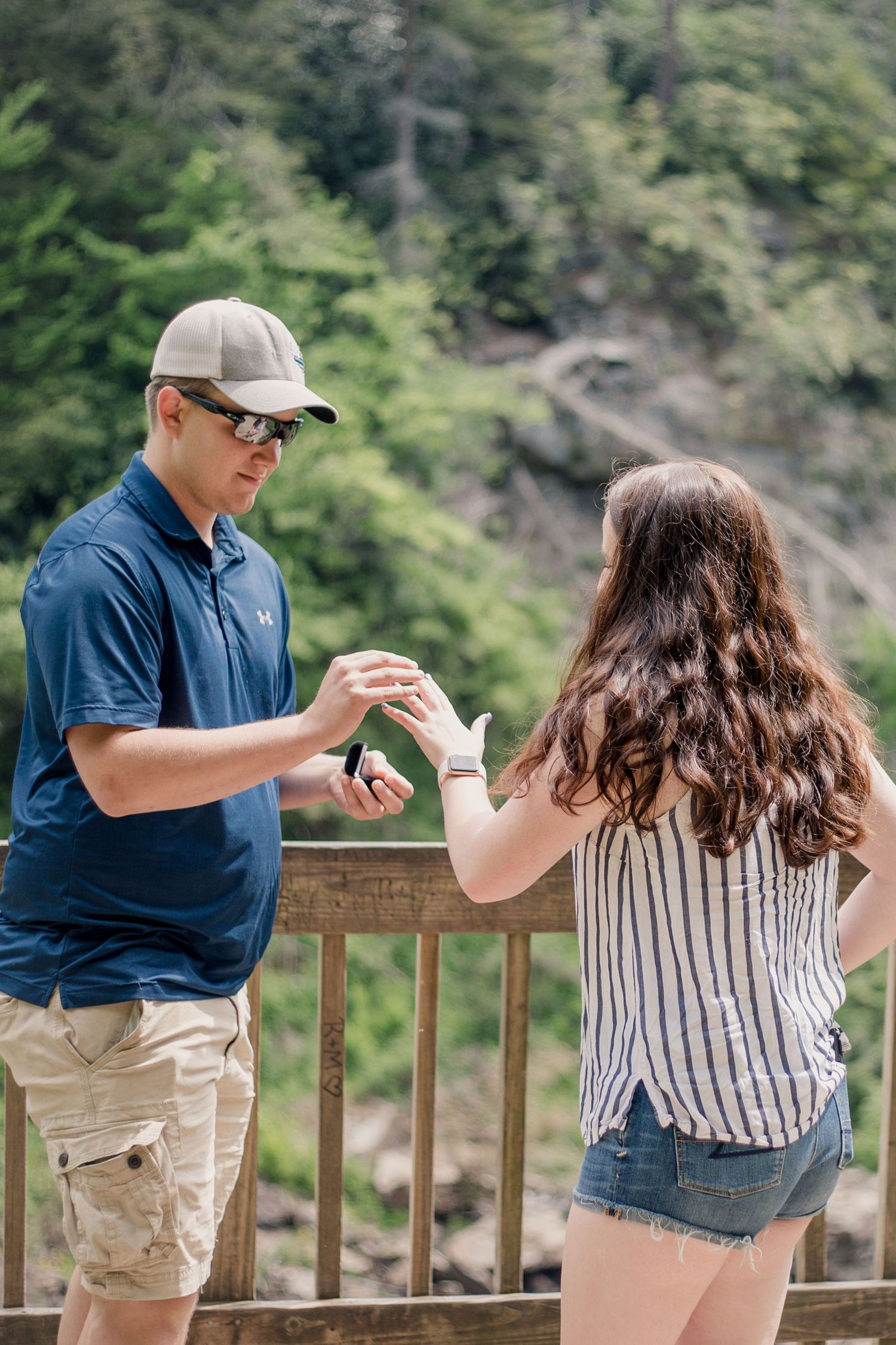 Two people standing on a viewing platform with lush greenery and mountains in the background.