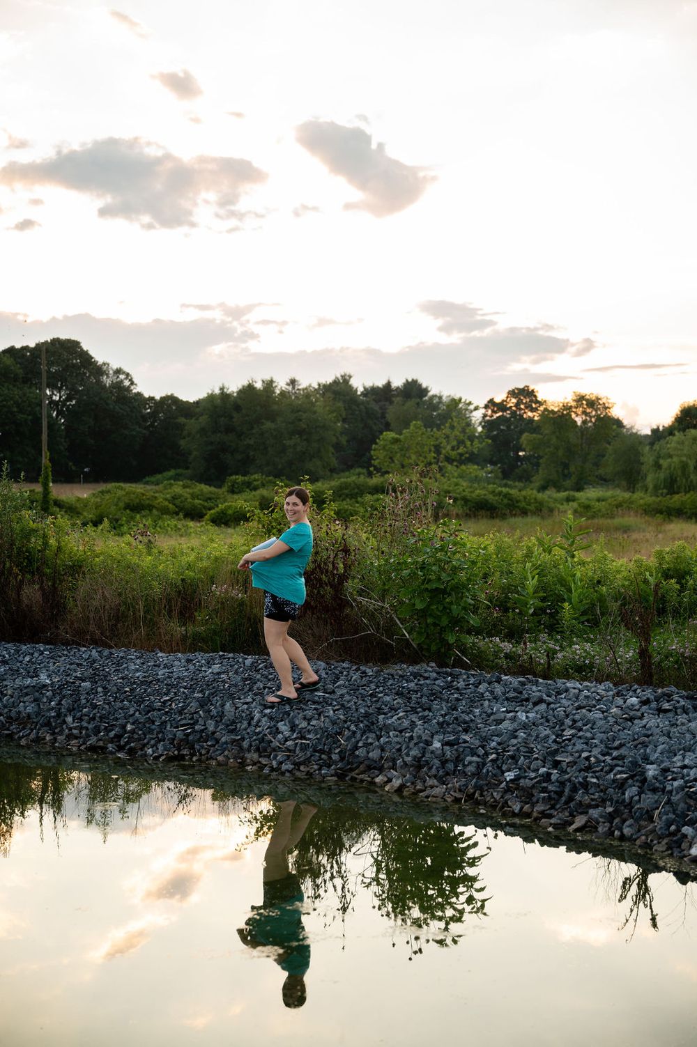 A person in blue stands on rocks by a pond at sunset with their reflection visible in the still water.
