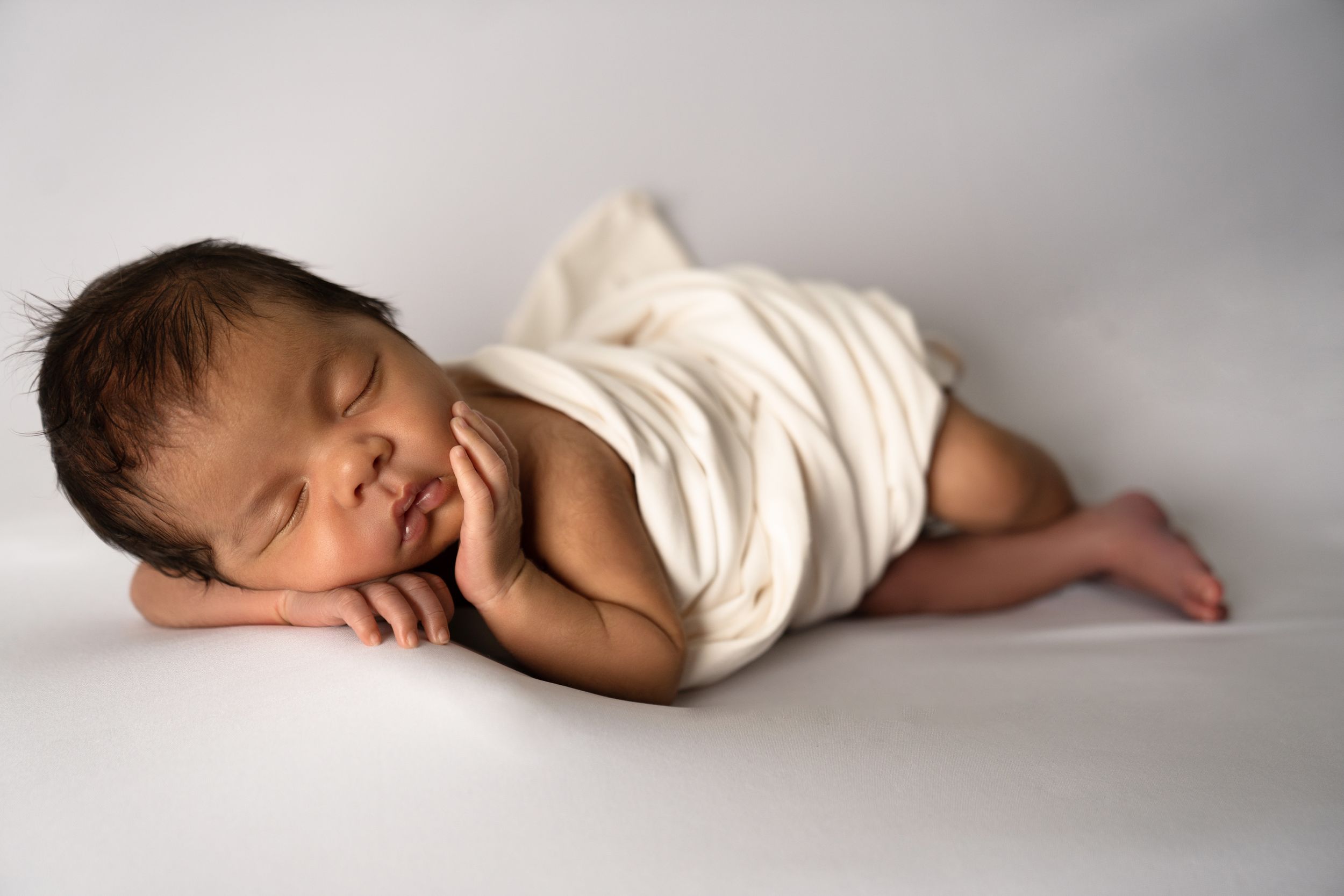 Peaceful sleeping newborn wrapped in white swaddle on light backdrop.