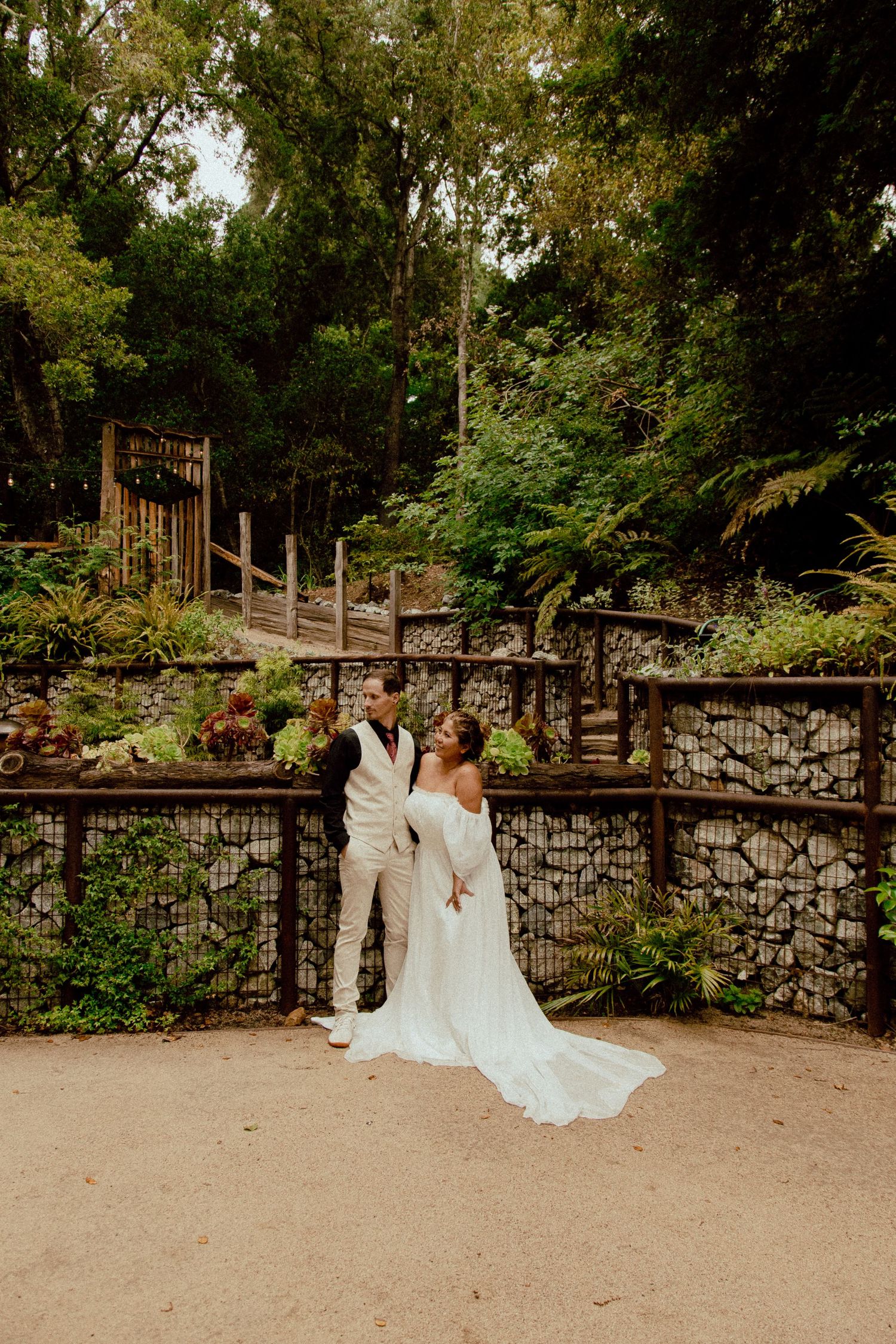 A couple shares a romantic moment on a stone path surrounded by lush greenery in a garden setting.