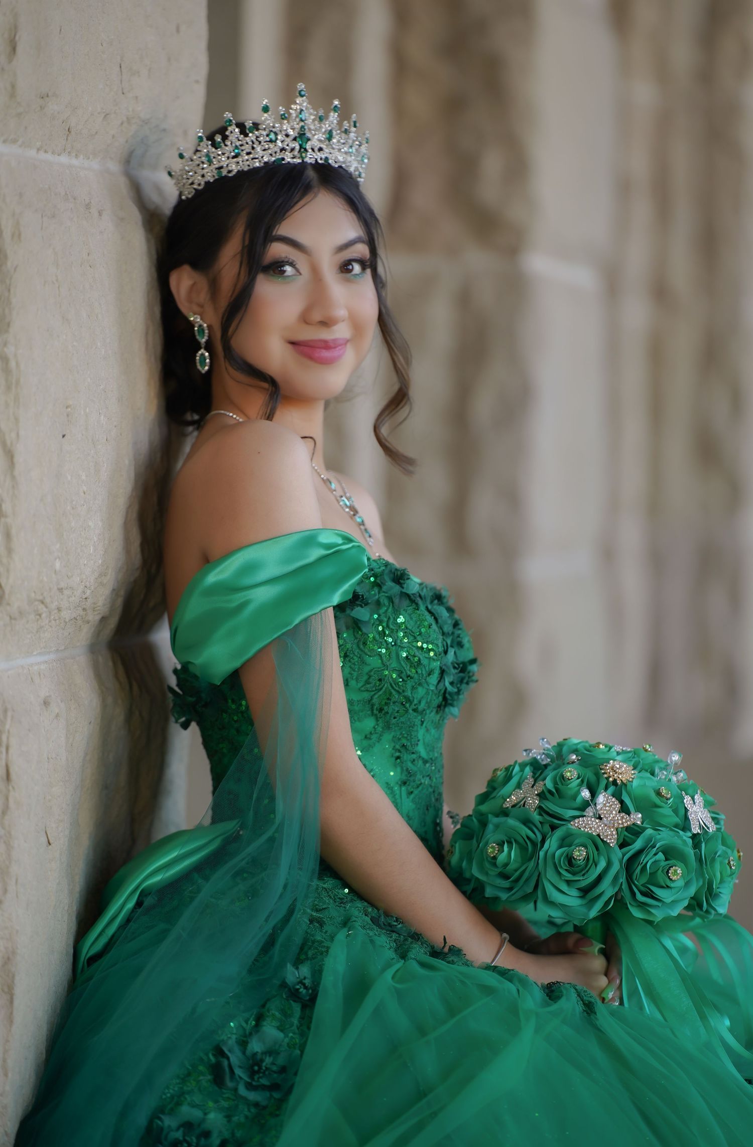 A person in a stunning emerald green ball gown and crown poses with matching floral bouquet.