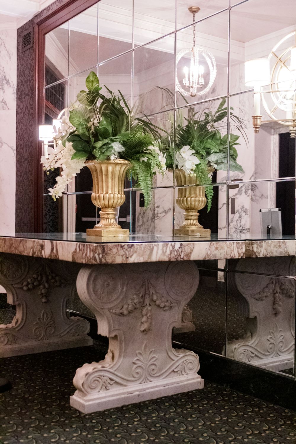 Elegant marble console table with ornate base displays gold urns filled with white floral arrangements.