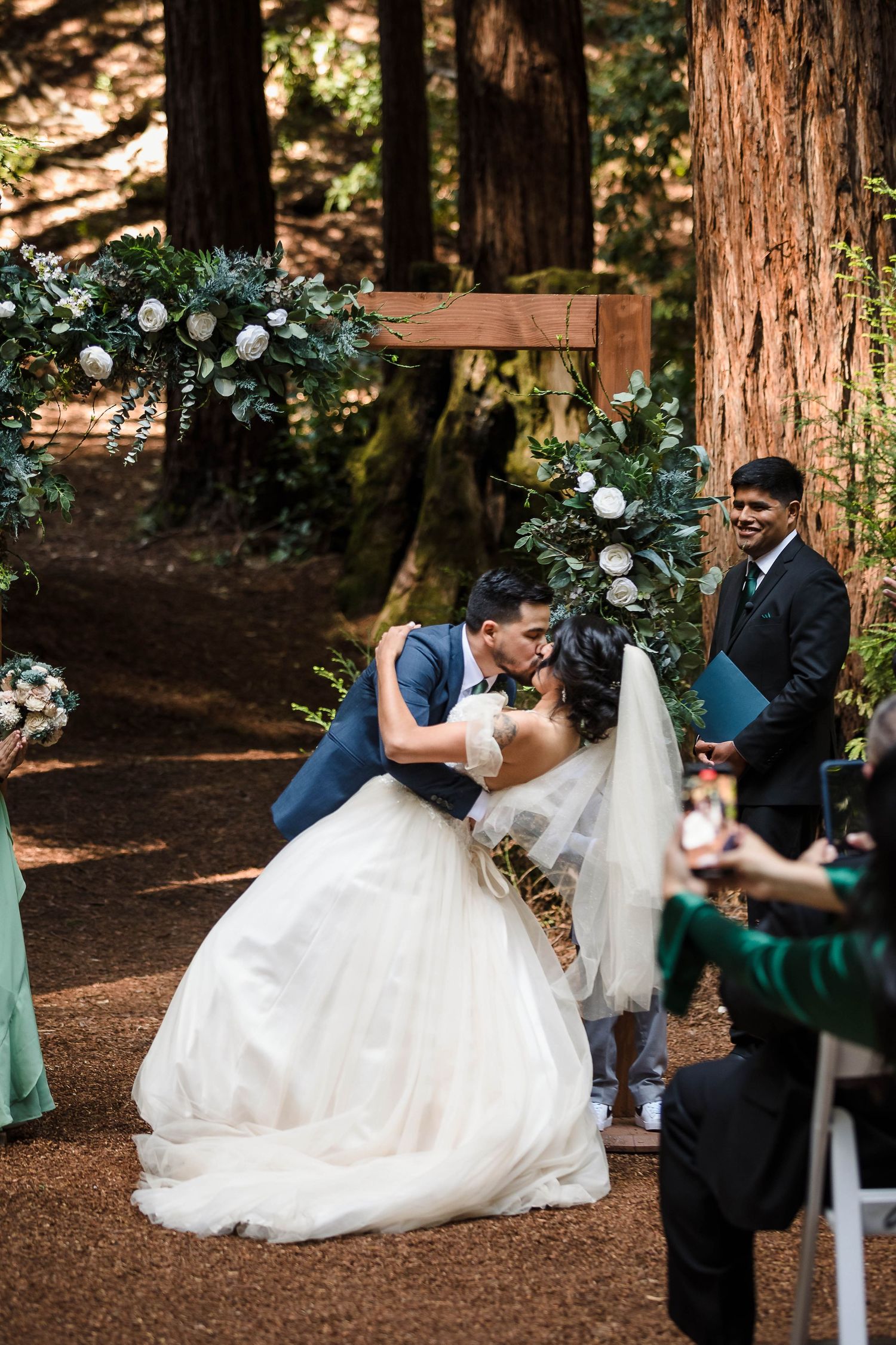 A romantic kiss shared beneath a floral arch in a majestic redwood forest setting.
