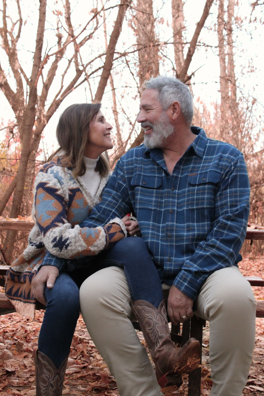 A couple wearing casual attire sits together on a bench surrounded by fall foliage.