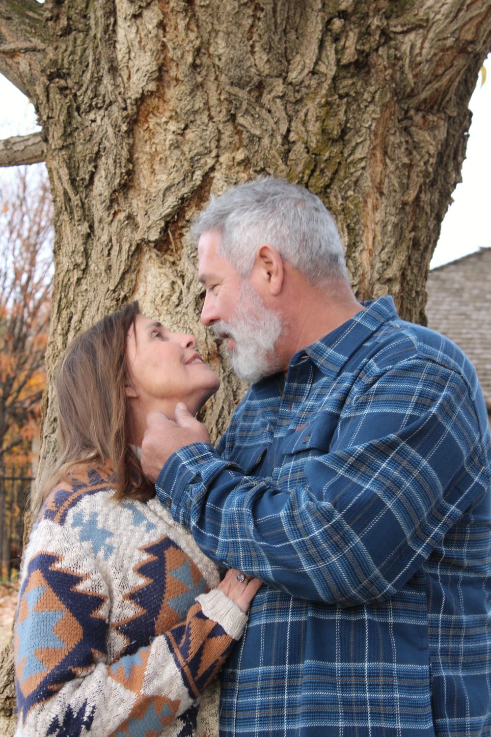 A couple in cozy fall sweaters share an intimate moment against a tree trunk in an autumn setting.