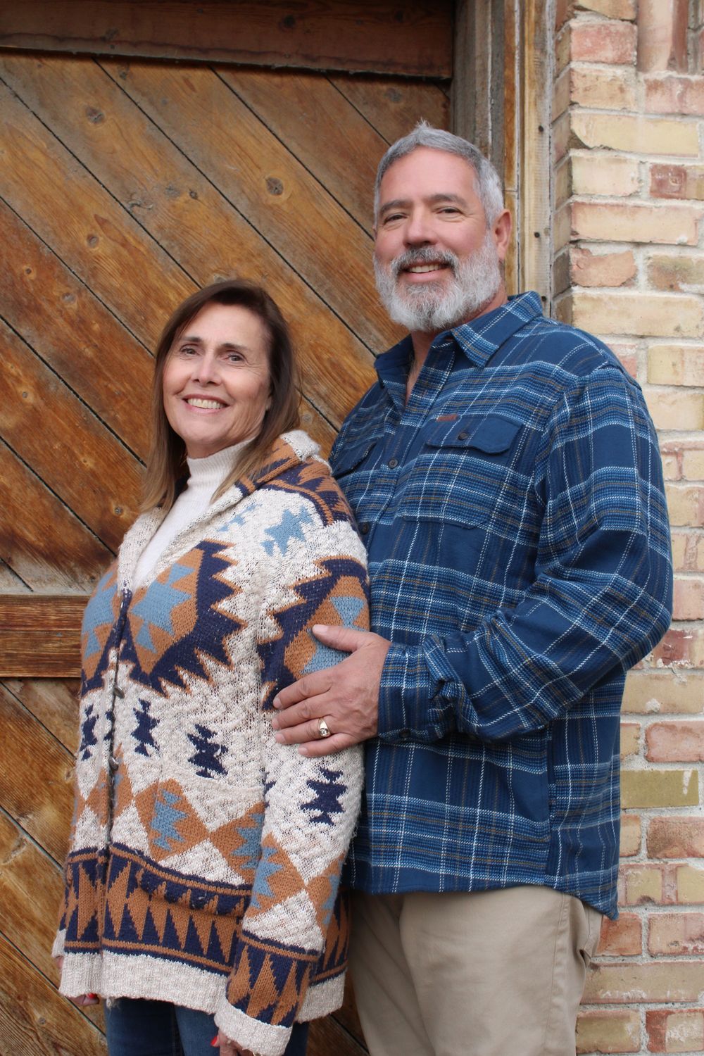 Couple poses against wooden and brick wall wearing casual fall attire.
