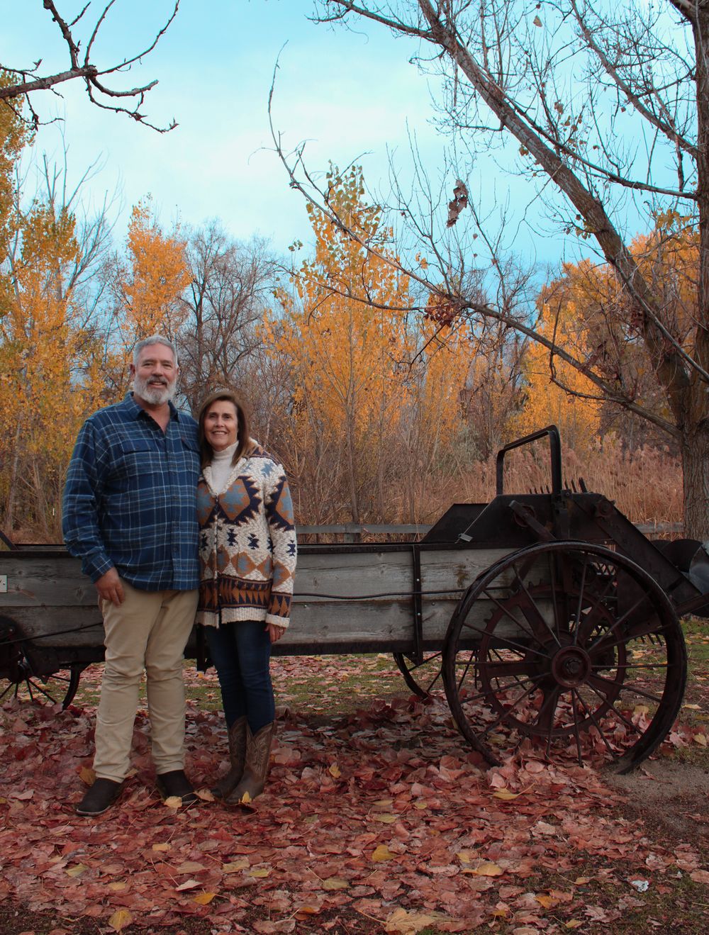 Couple stands by vintage farm equipment surrounded by fall foliage and fallen leaves.