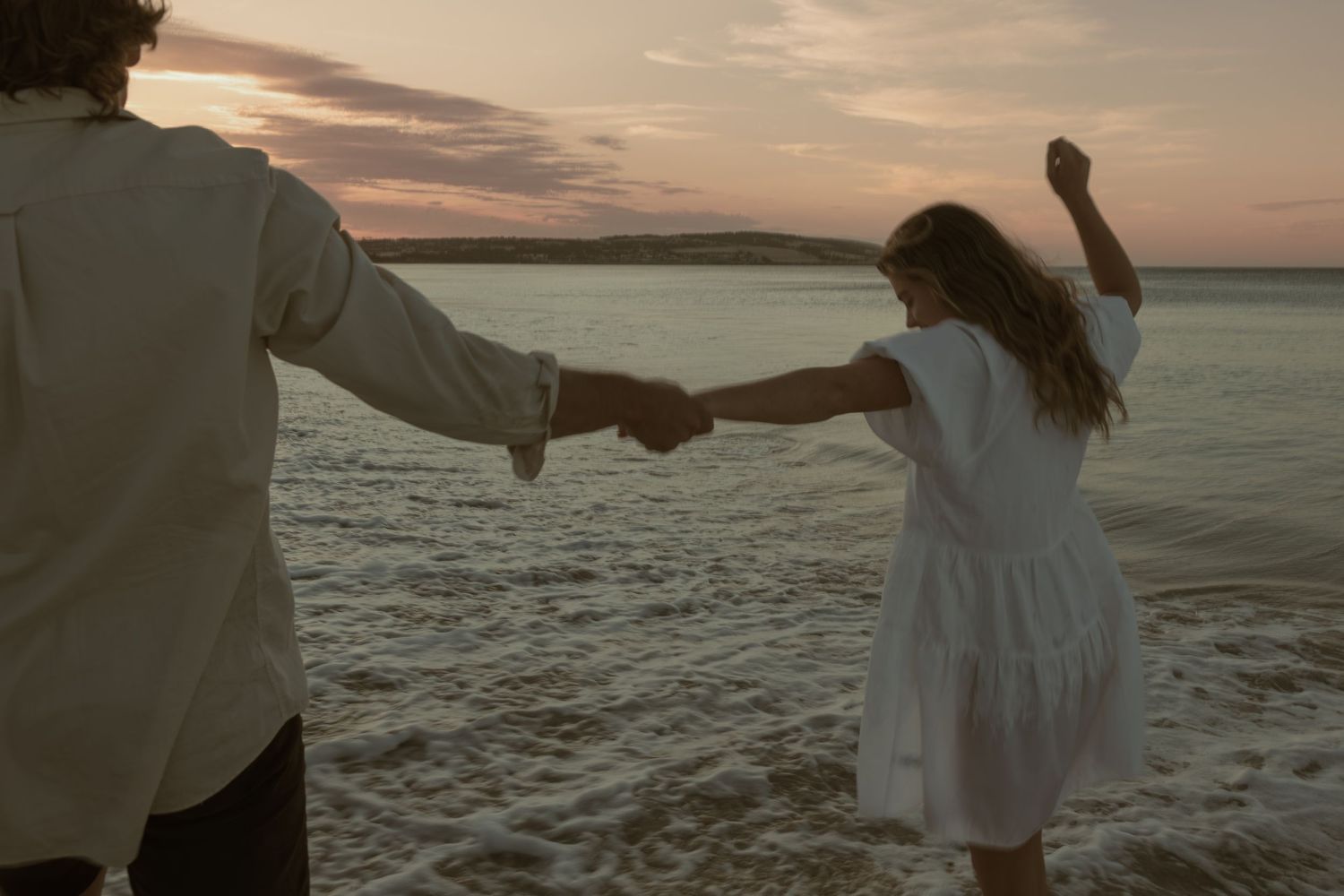 Couple walking hand in hand along a beach at sunset with water and cliffs in background.