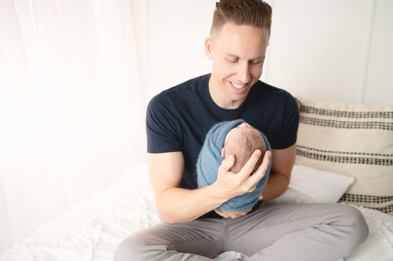 Parent in navy shirt holding and gazing lovingly at newborn on bed with striped pillows.