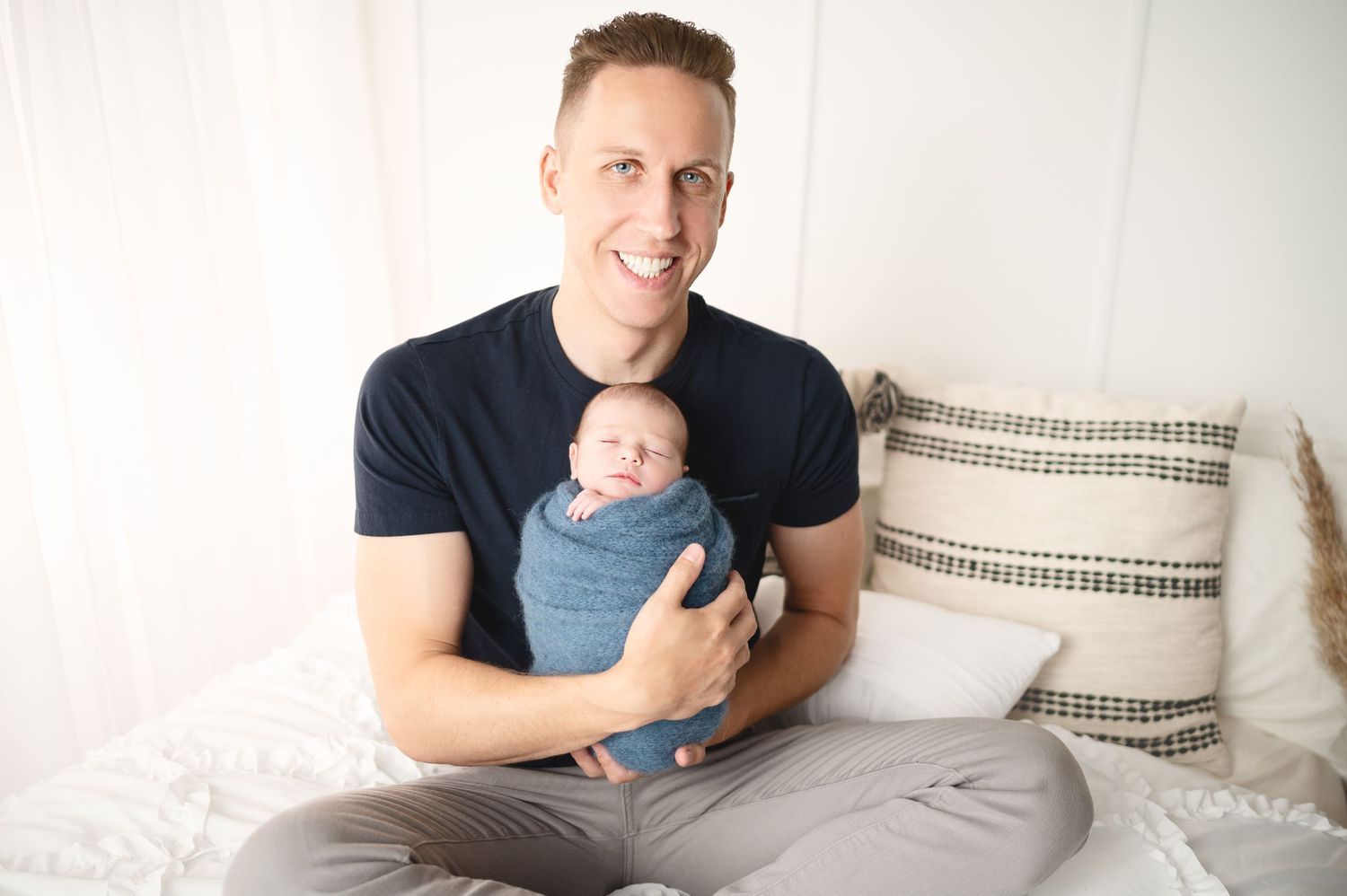 Parent in dark t-shirt holding small baby on bed with striped pillows.