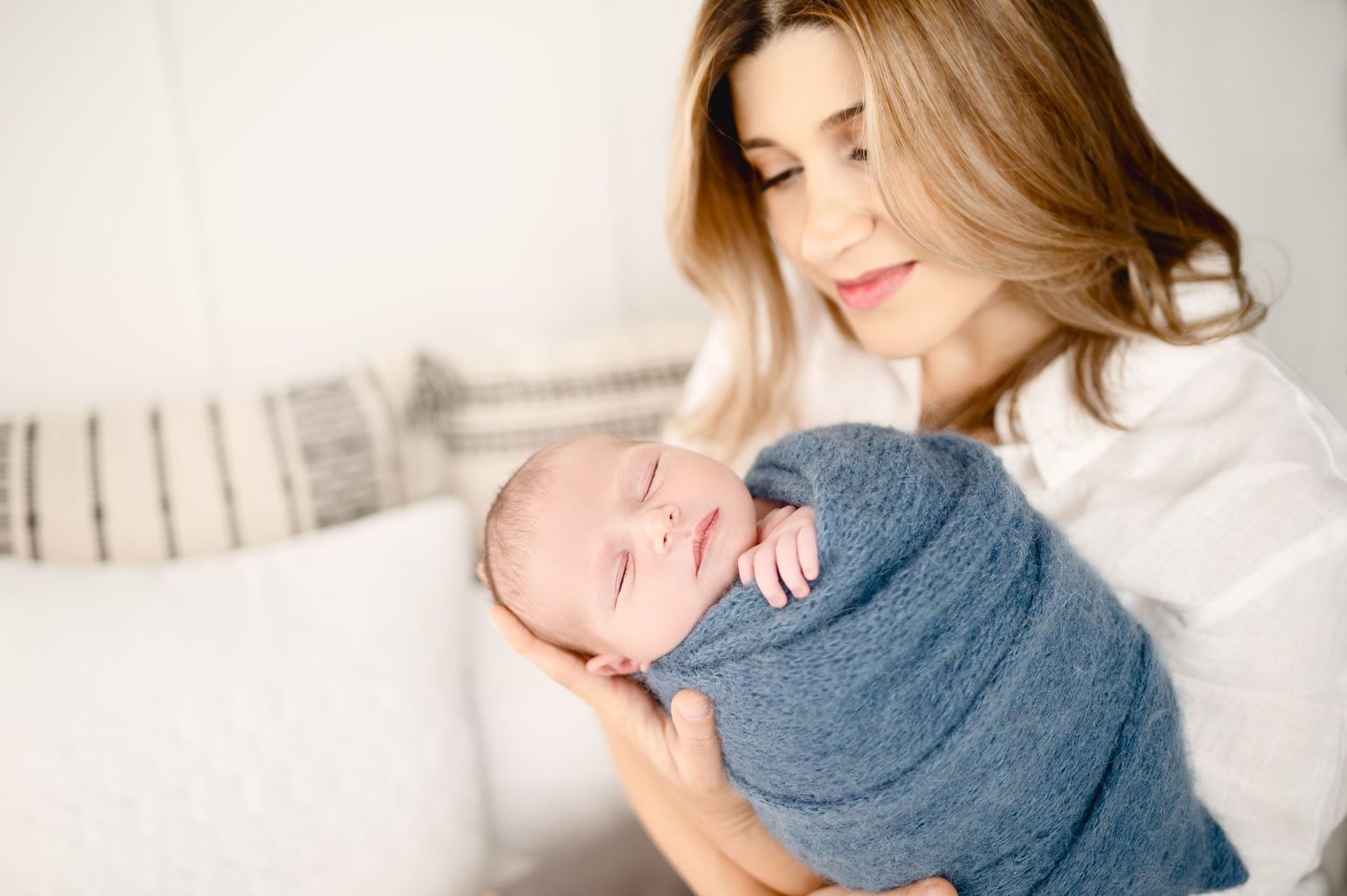 Mother in white sweater holding sleeping newborn wrapped in blue blanket.