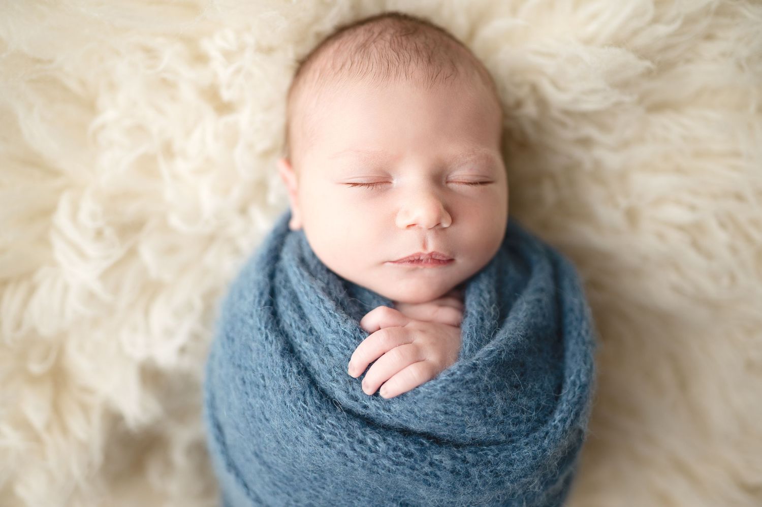 Sleeping newborn wrapped in cozy blue knit blanket on cream fur background.