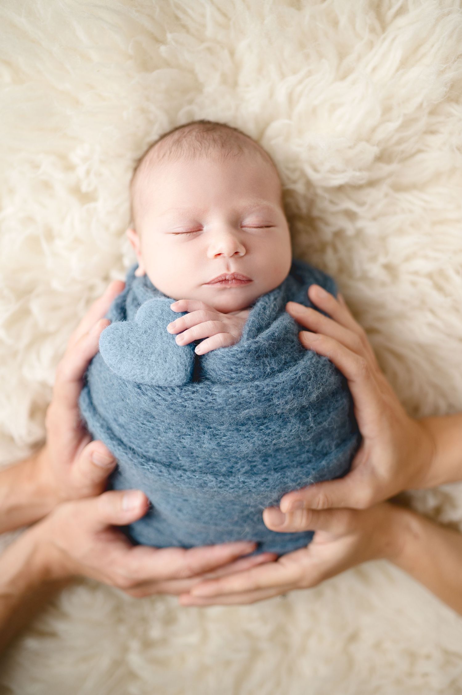Sleeping newborn wrapped in blue blanket on cream fur backdrop with gentle hands supporting.
