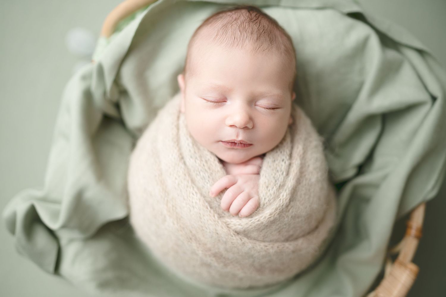 Sleeping newborn wrapped in beige swaddle on sage green backdrop in artistic composition.