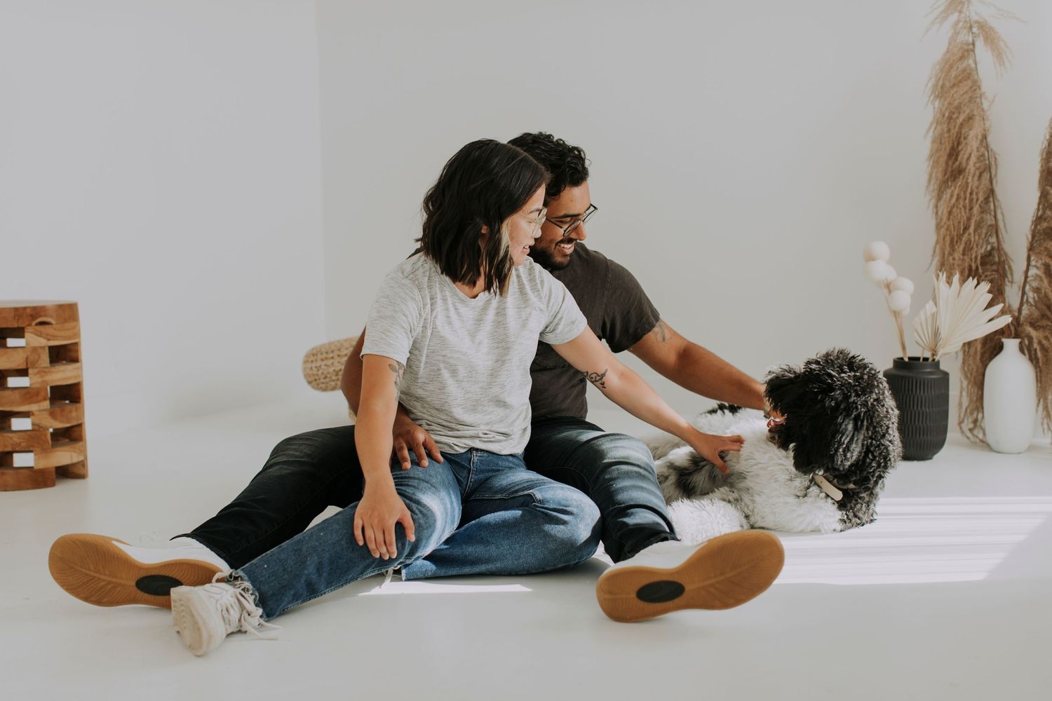 A couple sits together on a white floor playing with their small black dog.