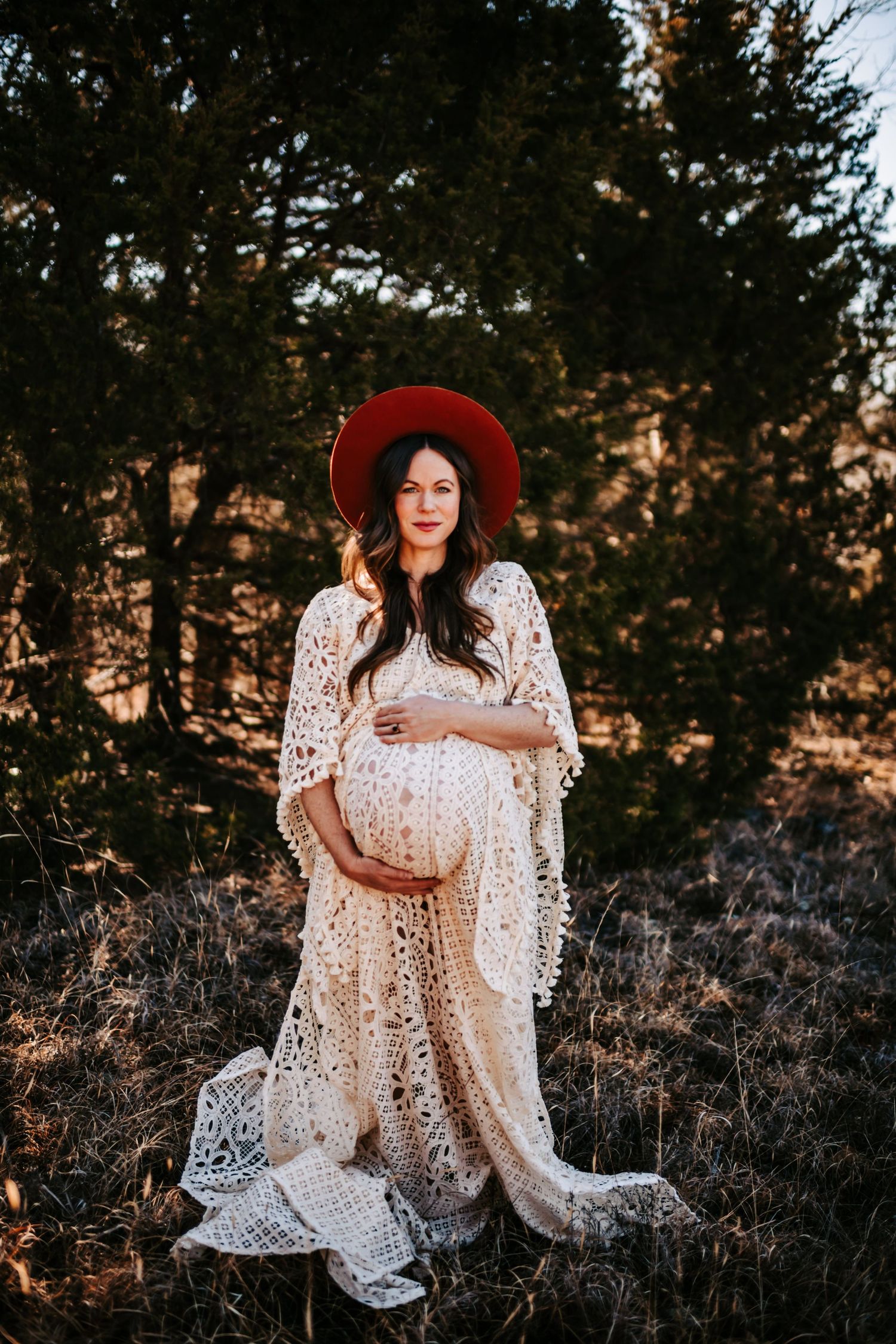 Maternity portrait in white lace dress and red hat during golden hour.