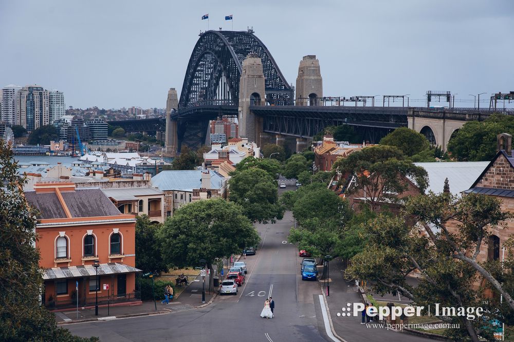 Bride in white gown walking down historic street with Sydney Harbour Bridge in background.