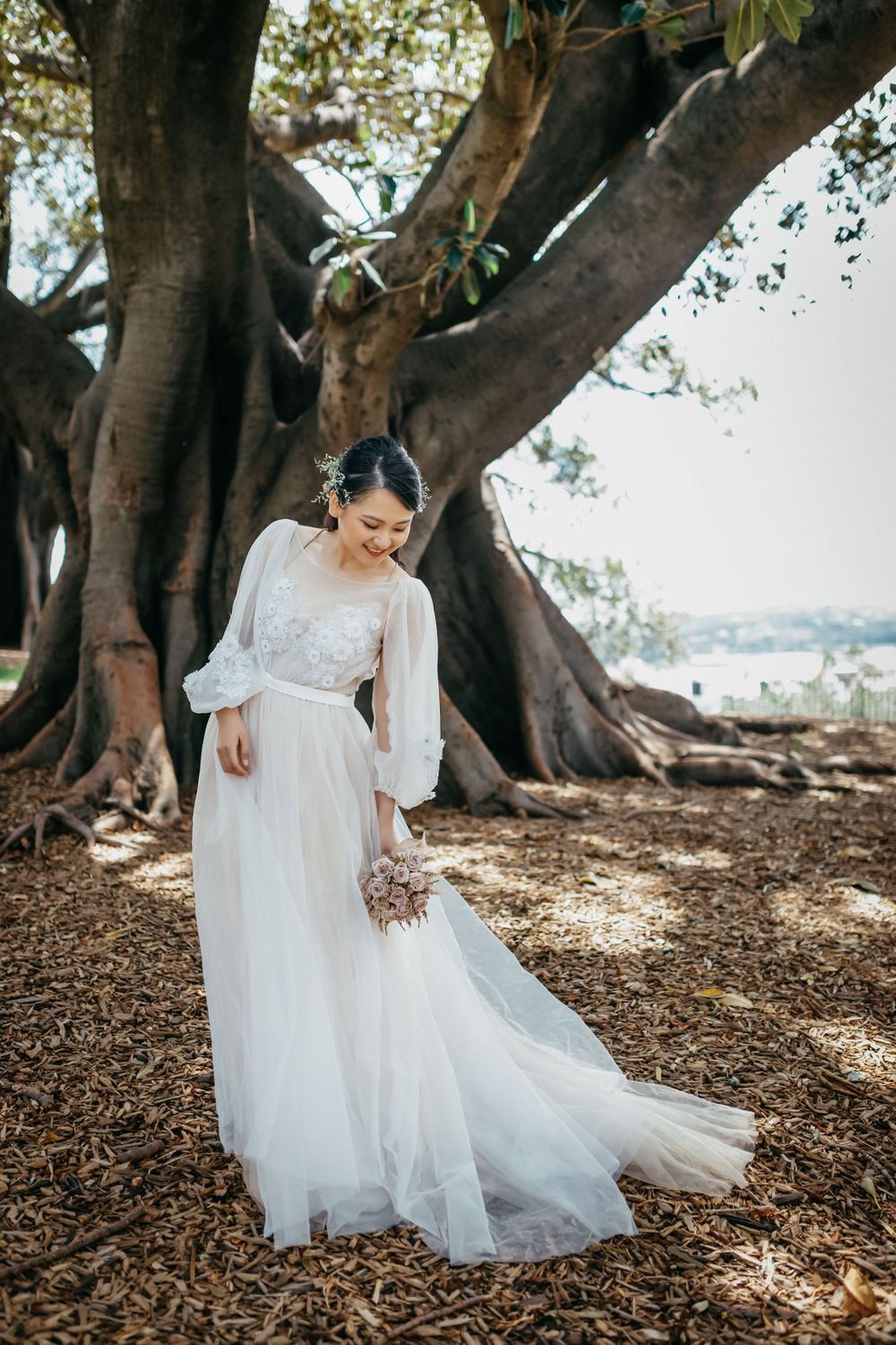 Bride in white lace gown holding bouquet standing under large fig tree in outdoor wedding portrait.
