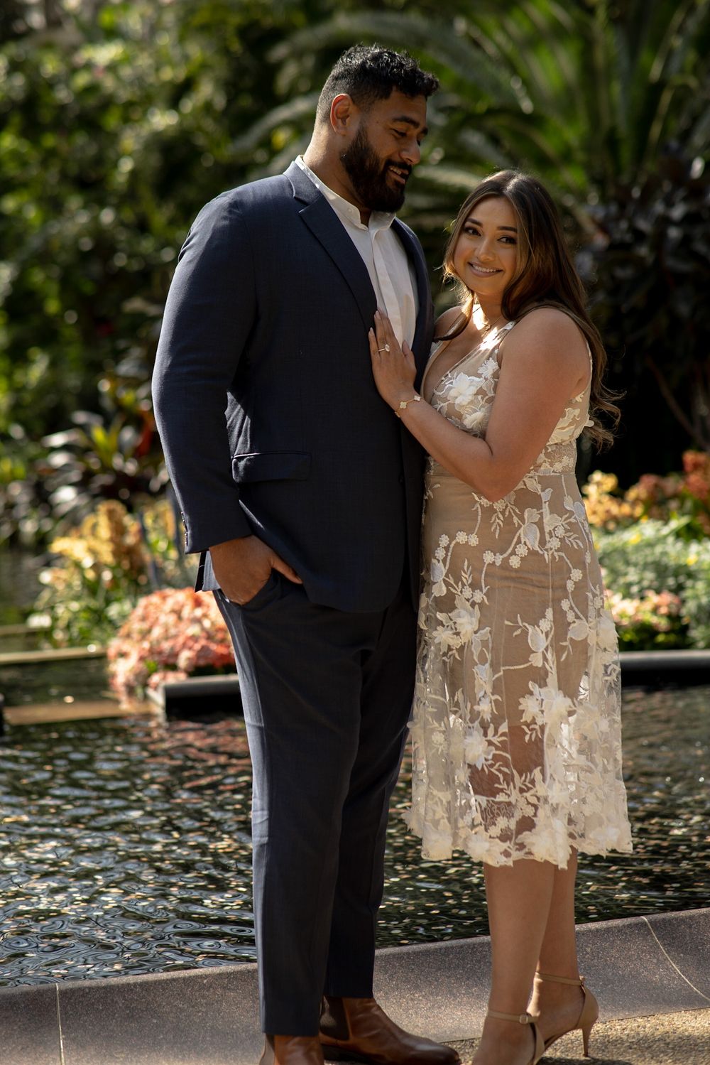 A couple poses elegantly by a garden fountain in formal wear.